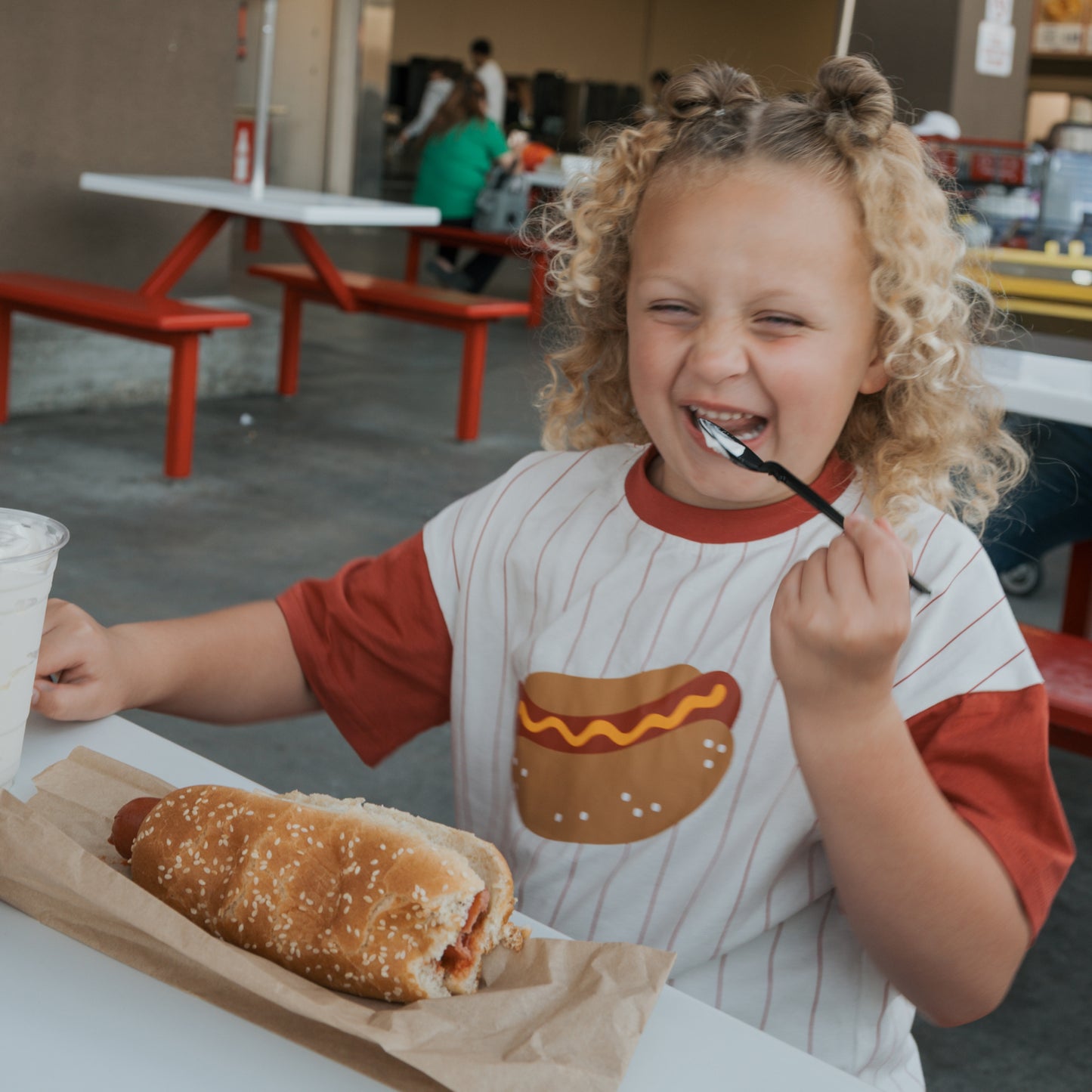 A happy child in the forever french baby Child Cotton Tee | Hot Dogs, featuring a playful cartoon hot dog, sits at a table ready to enjoy a hot dog and drink—making mealtime extra fun in their matching kids outfit.