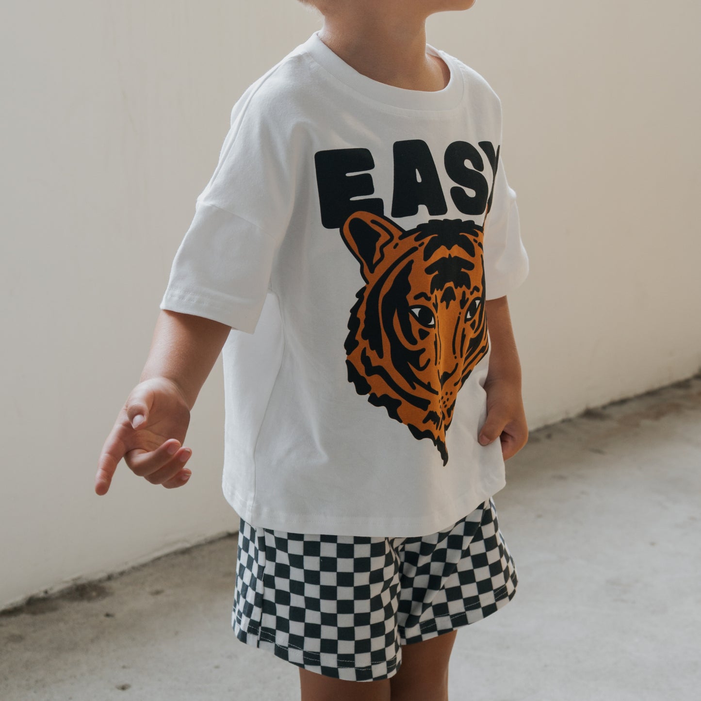 A young child wears the forever french baby Child Short Set | Easy Tiger—featuring a white T-shirt with a tiger graphic and "EASY," paired with black and white checkered shorts—standing against a light background.