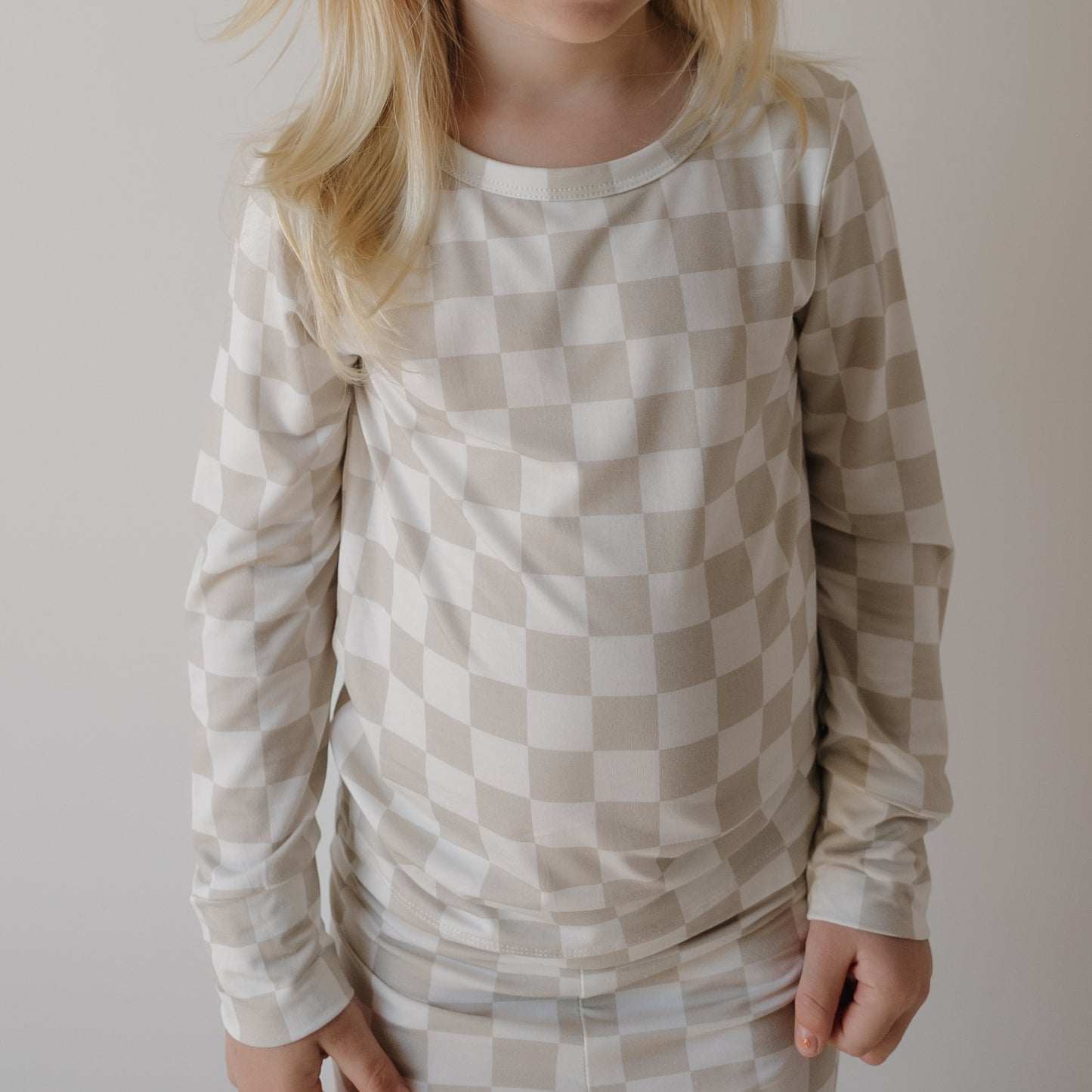 A young child in the forever french baby Child Sleepy Time Set | Checkerboard—cream and beige checks made from soft, hypo-allergenic materials—stands against a pale background, face partly out of frame.