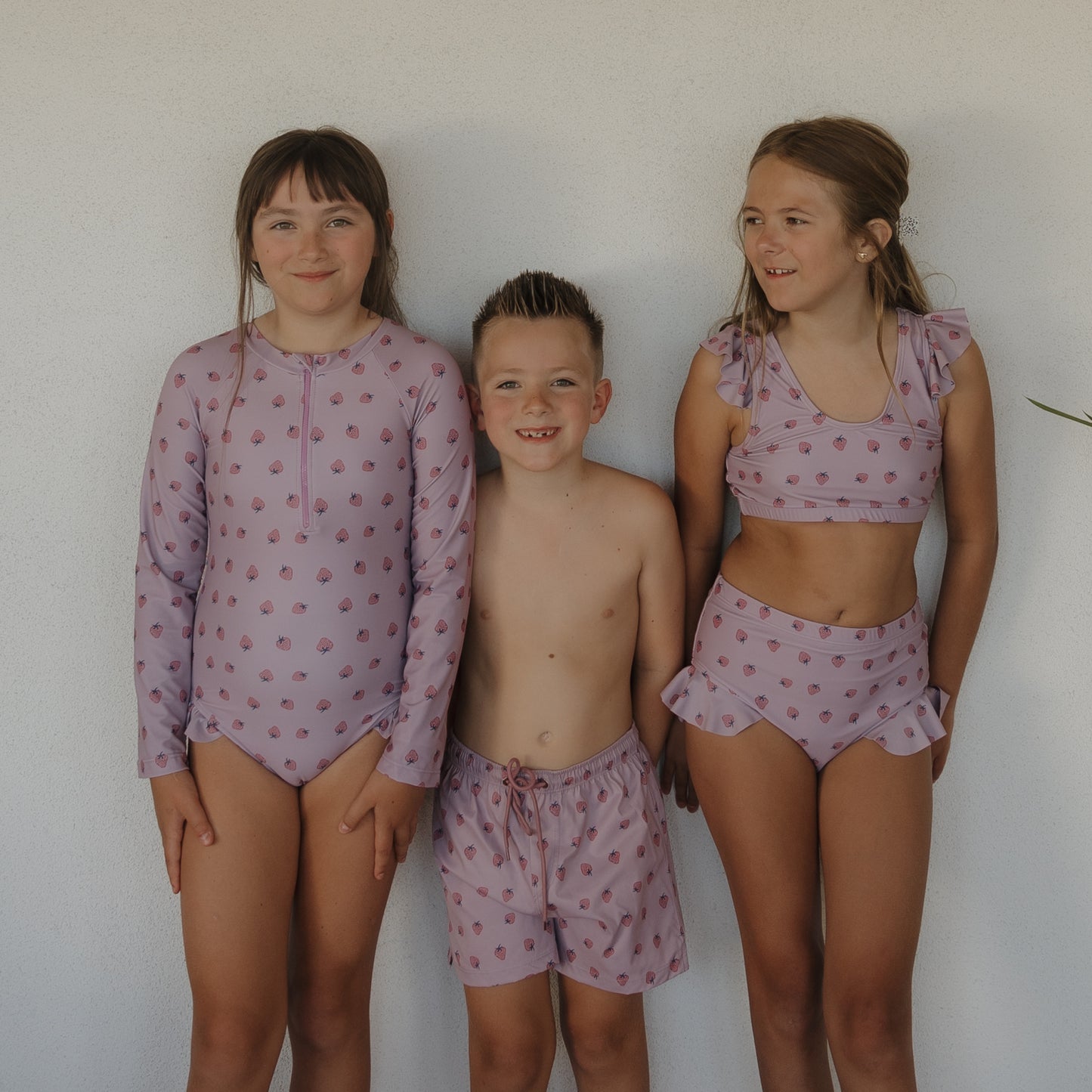 Two girls and a boy stand against a white wall. The girl on the right wears the forever french baby Girl’s Two Piece Swimsuit | Matilda, while the others wear coordinating kids swimwear.