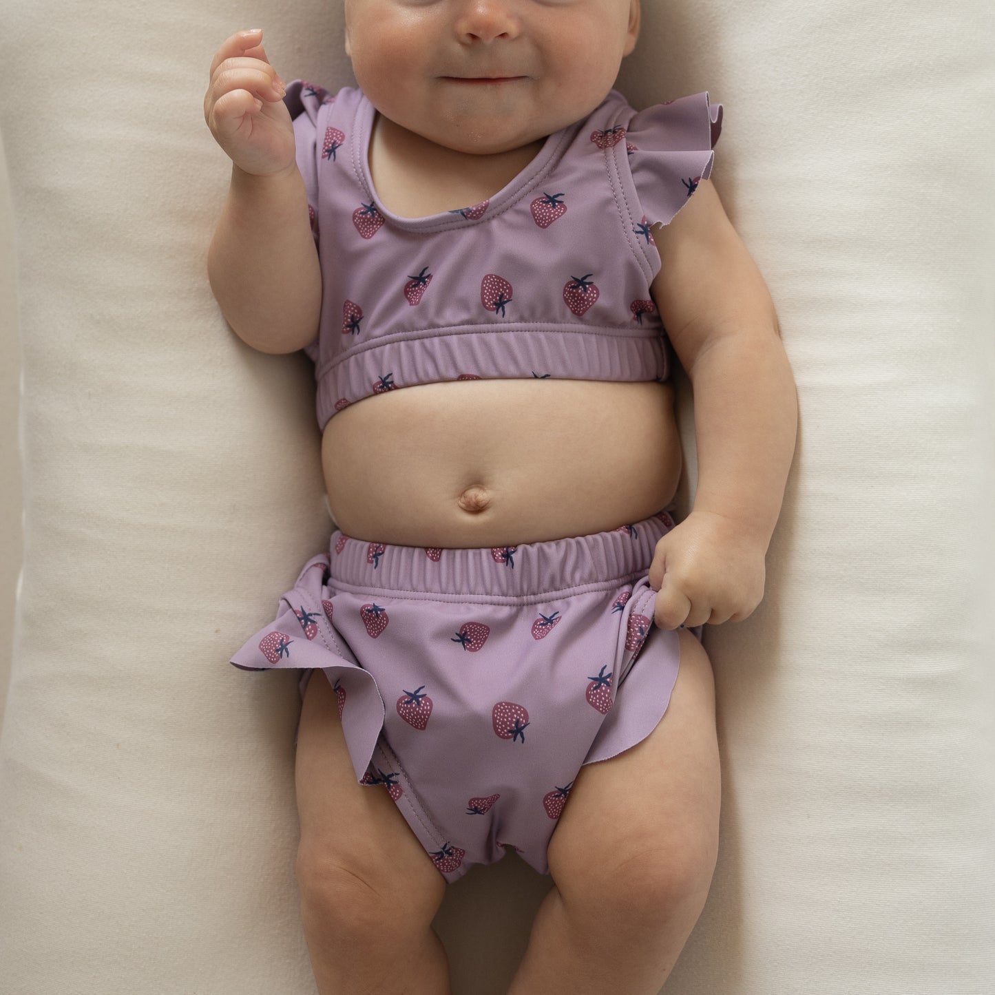 A baby smiles softly while wearing the forever french baby Girl's Two Piece Swimsuit | Matilda, featuring a lavender hue, ruffled sleeves, and pink strawberry print—a sweet and stylish choice for little swimmers.