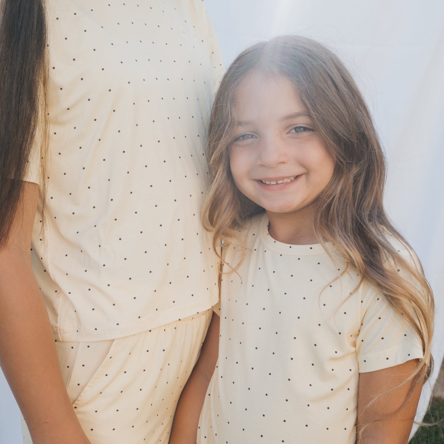 A young girl with long, wavy hair smiles beside an adult as they both wear matching "Bamboo Short Set | Veda" pajamas by forever french baby—cream-colored with black polka dots, perfect for breathable children's clothing.