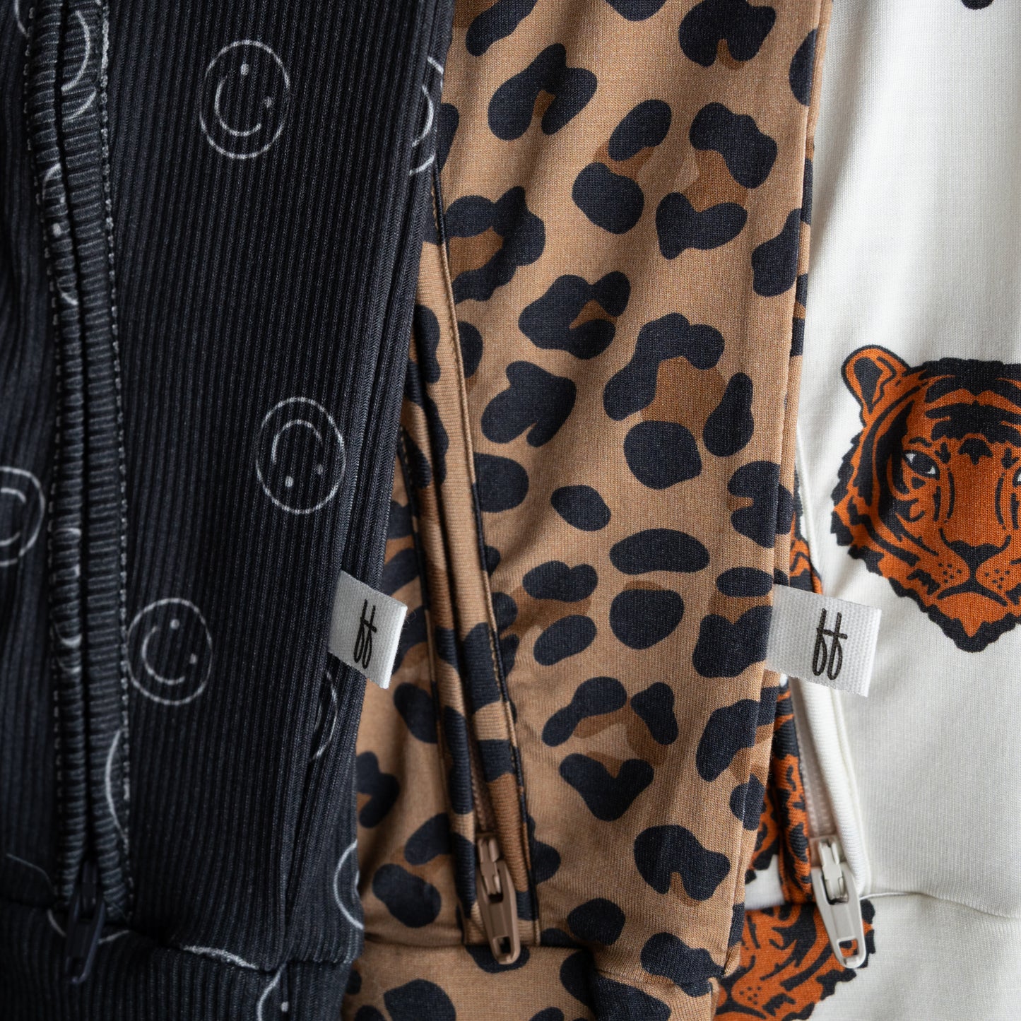 Close-up of three forever french baby Bamboo Zip Pajamas: black with white smiley faces, brown with black leopard spots, and white with a tiger face. Each features soft, breathable fabric and a small white "ff" tag near the zipper.