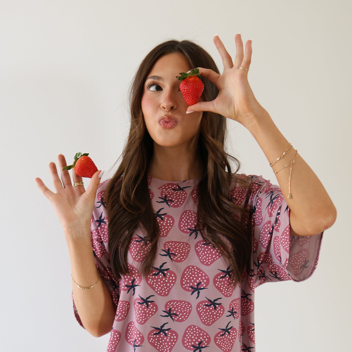 A woman with long brown hair wears the forever french baby Women's Bamboo Short Set | Matilda, featuring a pink strawberry pattern. She playfully holds two strawberries—one near her mouth, one by her eye—against a plain white background.