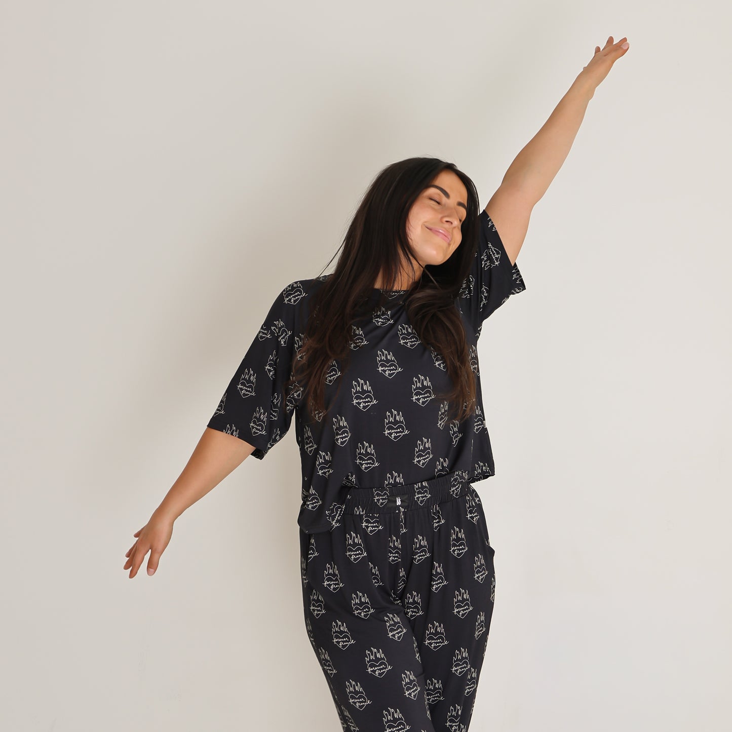 A woman wears the Women's Boxy Tee Lounge Set in Flaming Hearts by forever french baby, standing against a plain white background with her eyes closed and arms stretched out, looking joyful and relaxed.