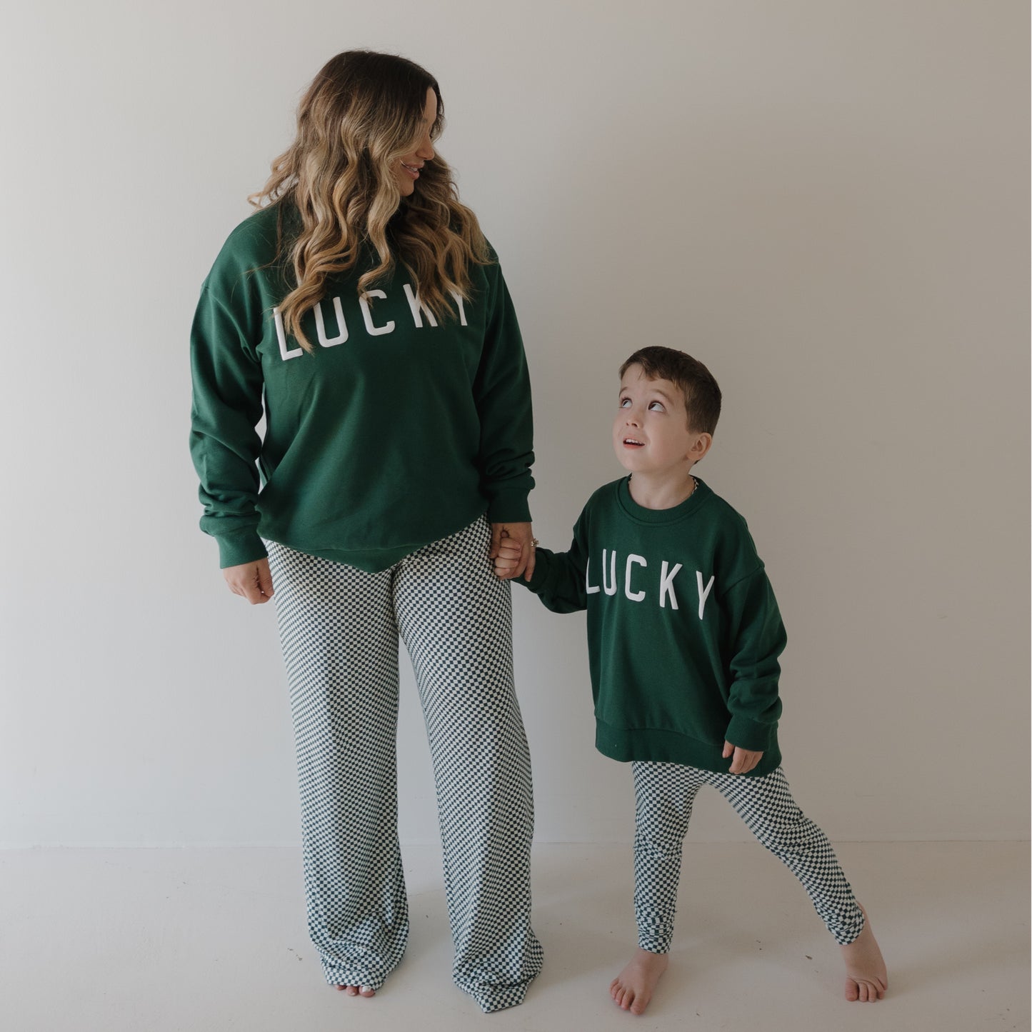 A woman and young boy smile barefoot against a white wall, holding hands in matching forever french baby "Women's Boxy Tee Lounge Set | Lucky Micro Check," enjoying the soft, hypo-allergenic comfort of their bamboo outfits.