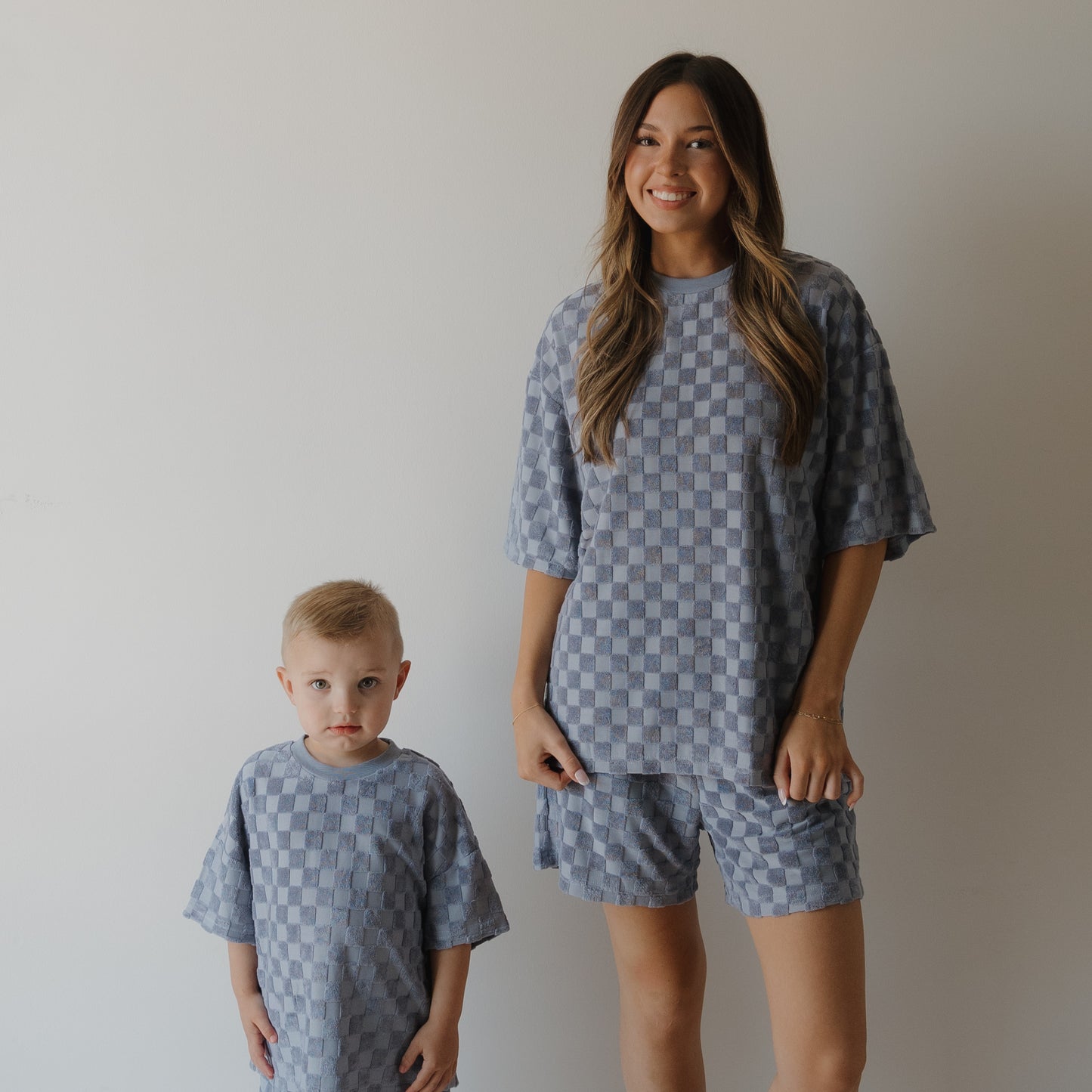 A woman and a young boy stand side by side in blue checkerboard outfits from forever french baby, both wearing the Women's Terry Short Set | Tradewinds, against a white background. The woman smiles as the boy looks at the camera.