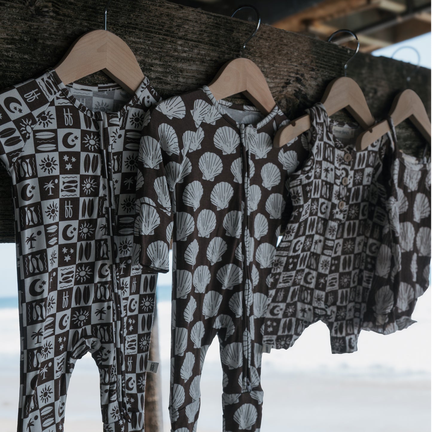Four forever french baby Bamboo Zip Pajamas | Seashells hang on wooden hangers from a rustic beam, with a blurred beach and ocean backdrop—perfect breathable sleepwear for little ones.