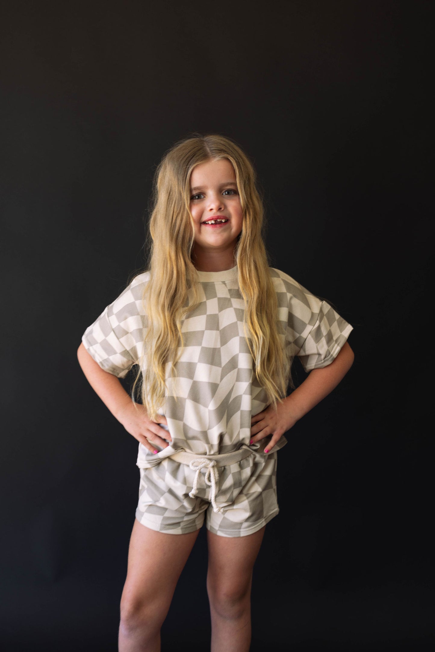 A young girl with long blonde hair stands confidently with her hands on her hips against a black background. She is wearing a trendy Short Set | Wavy Checkerboard from the brand forever french baby, crafted from organic cotton and showcasing light and dark shades of beige in a stylish wavy checker pattern.