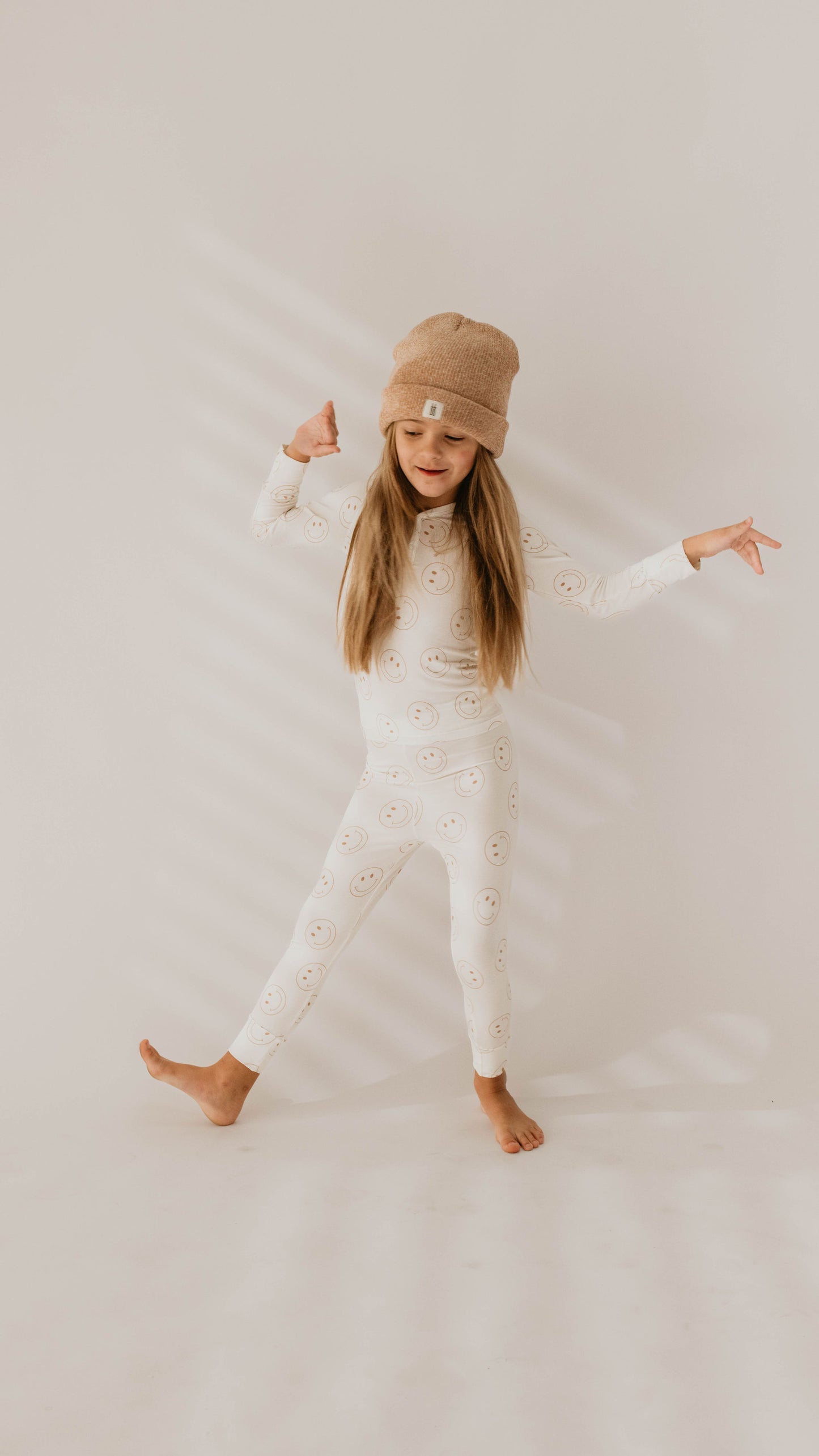 A young girl with long hair stands barefoot on a light-colored floor against a plain background. She is wearing a beige beanie and a matching white outfit made of breathable Bamboo Two Piece Pajamas from the "Just Smile" collection by forever french baby, featuring light pink smiley faces. She poses with her arms extended and one foot lifted, appearing playful and joyful.