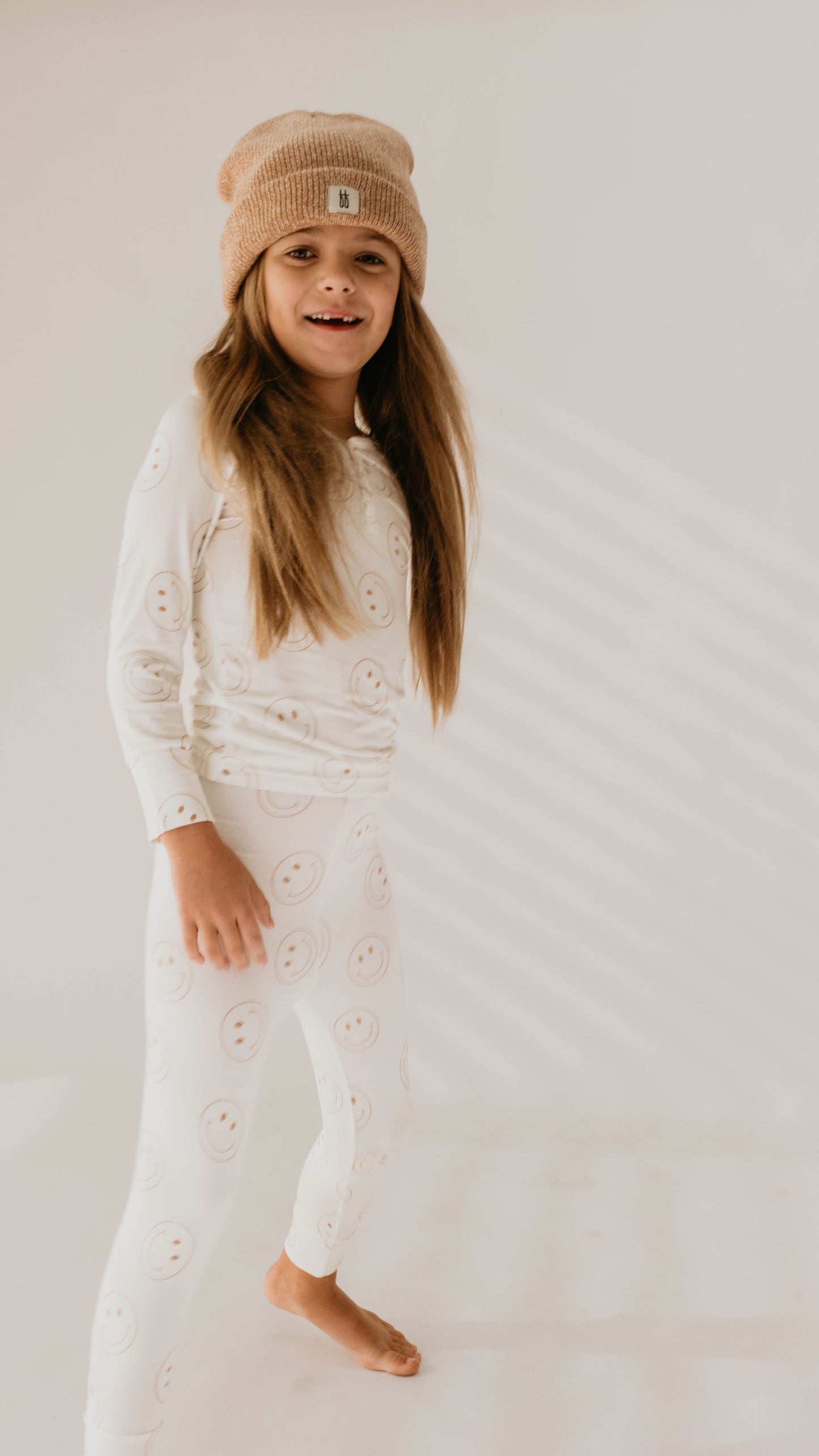 A happy child with long hair, clad in a beige beanie and matching Bamboo Two Piece Pajamas | Just Smile from forever french baby, stands barefoot on a light background. The breathable baby clothes feature cheerful smiley face designs, and the child gazes slightly to the side as soft shadows cast behind them.