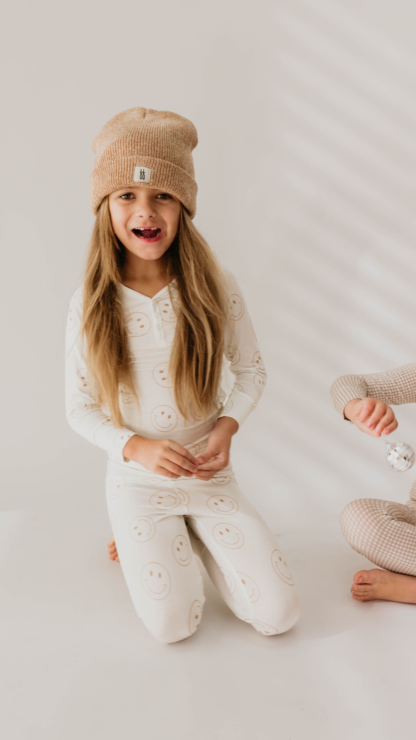 Two children dressed in cozy, hypo-allergenic clothing and knit hats. One child with long hair smiles while kneeling, wearing the "Bamboo Two Piece Pajamas | Just Smile" set in white with smiley faces by forever french baby. The other child sits cross-legged, partially out of view, in a checkered beige set made of breathable bamboo pajamas.