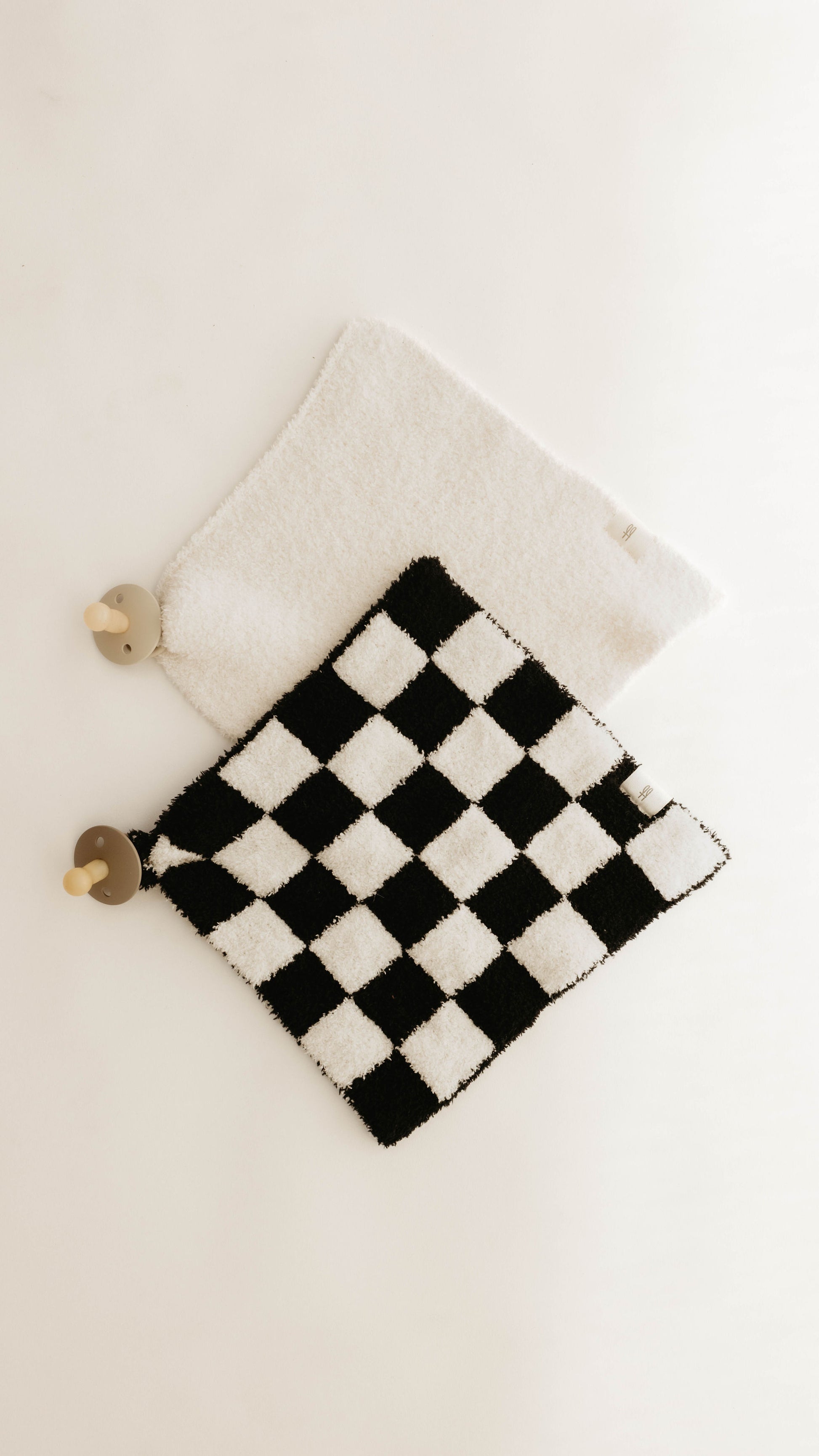 Two textured baby blankets are shown on a white background. One blanket, made from soft microfiber feather yarn, features a black and white checkered pattern, while the other is Cream. Each forever french baby Lovey has a round, plush animal head attached to one corner, resembling a rabbit and a bear.
