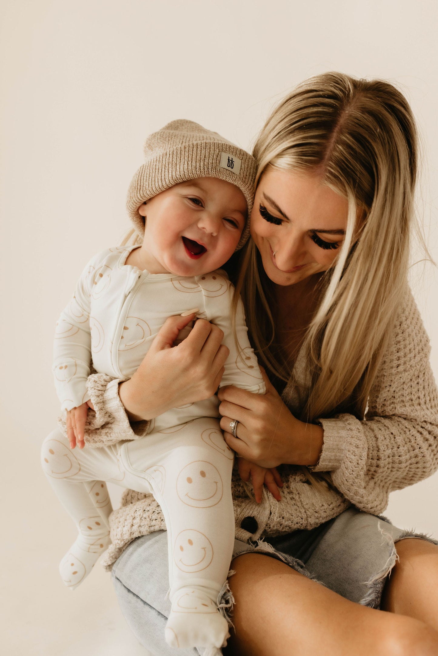 A woman with long blonde hair is sitting and smiling, holding a laughing baby in her lap. Both are wearing beige outfits; the baby is dressed in forever french baby's Bamboo Zip Pajamas | Just Smile with a matching beanie. The woman wears a knitted sweater and denim shorts. The background is plain.