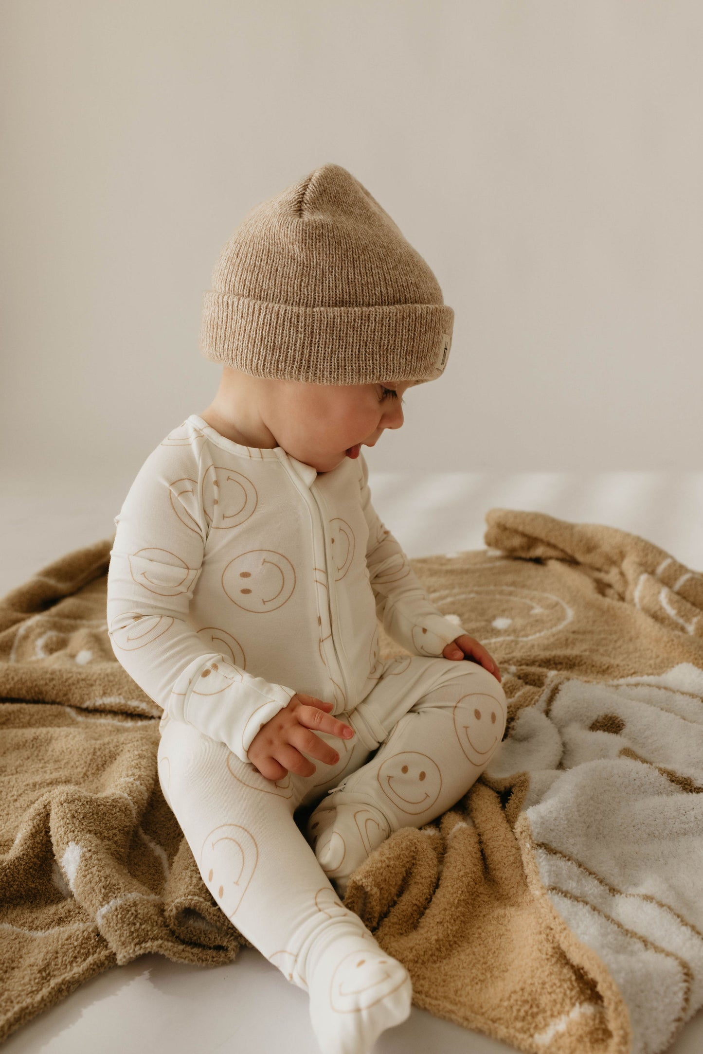 A baby, dressed in forever french baby's Bamboo Zip Pajamas | Just Smile, along with a beige knit hat and a white onesie adorned with smiley face patterns, sits on a beige blanket that also features a similar smiley face design. The baby looks down and seems to be playing with the blanket against a plain, light-colored background.