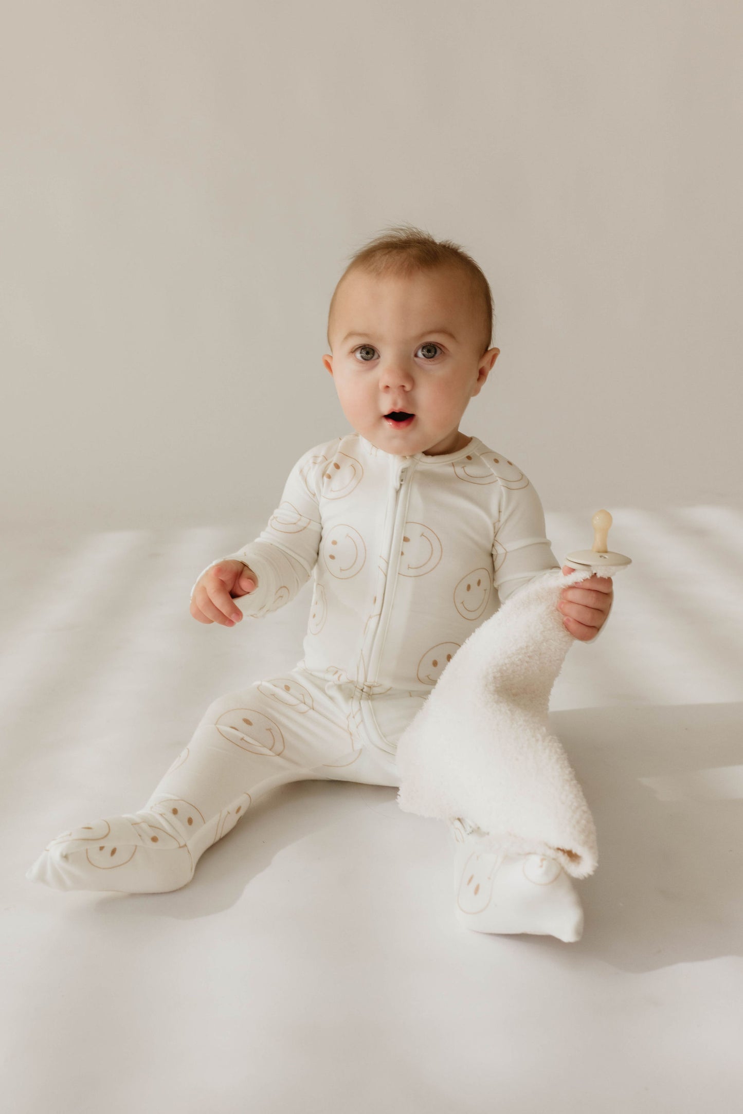 A baby in the forever french baby Bamboo Zip Pajamas | Just Smile, featuring cute smiley face prints, sits on a light-colored surface. The baby holds a soft white blanket and a pacifier, wearing hypo-allergenic clothing with an adorably curious expression, wide eyes, and slightly parted lips. Natural light softly illuminates the scene.