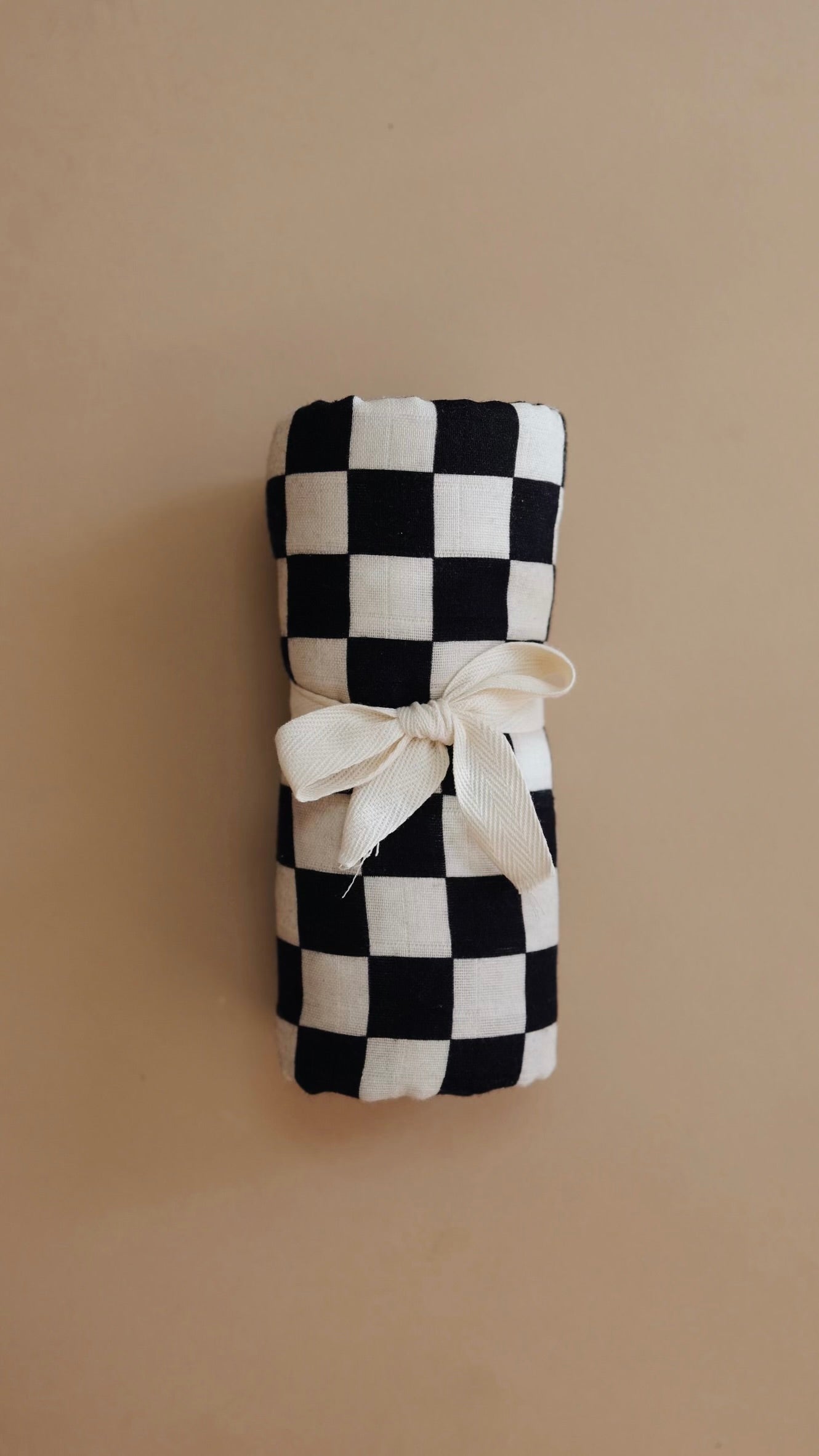 A forever french baby Muslin Swaddle in Black & White Checkerboard, rolled up and tied with a cream-colored ribbon, rests against a beige background.