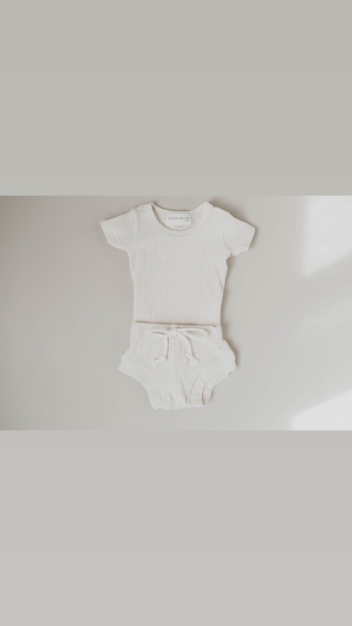 A gentle cream-hued Ribbed Set - Vanilla by forever french baby rests on a light surface, featuring an oversized ribbed cotton short-sleeve tee and matching shorts with a waistband tie. This adorable two-piece set offers comfort and style for your little one.