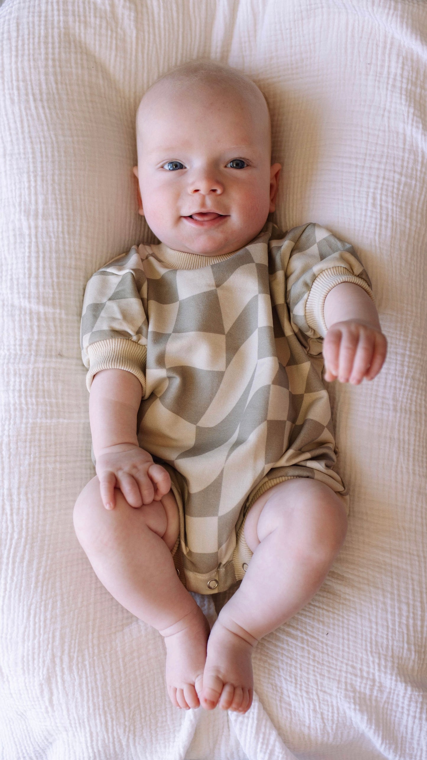 A baby wearing the "Short Sleeve Romper | Wavy Checkerboard" by forever french baby lies on a white textured blanket. The baby is looking up while smiling, with arms and legs slightly raised.