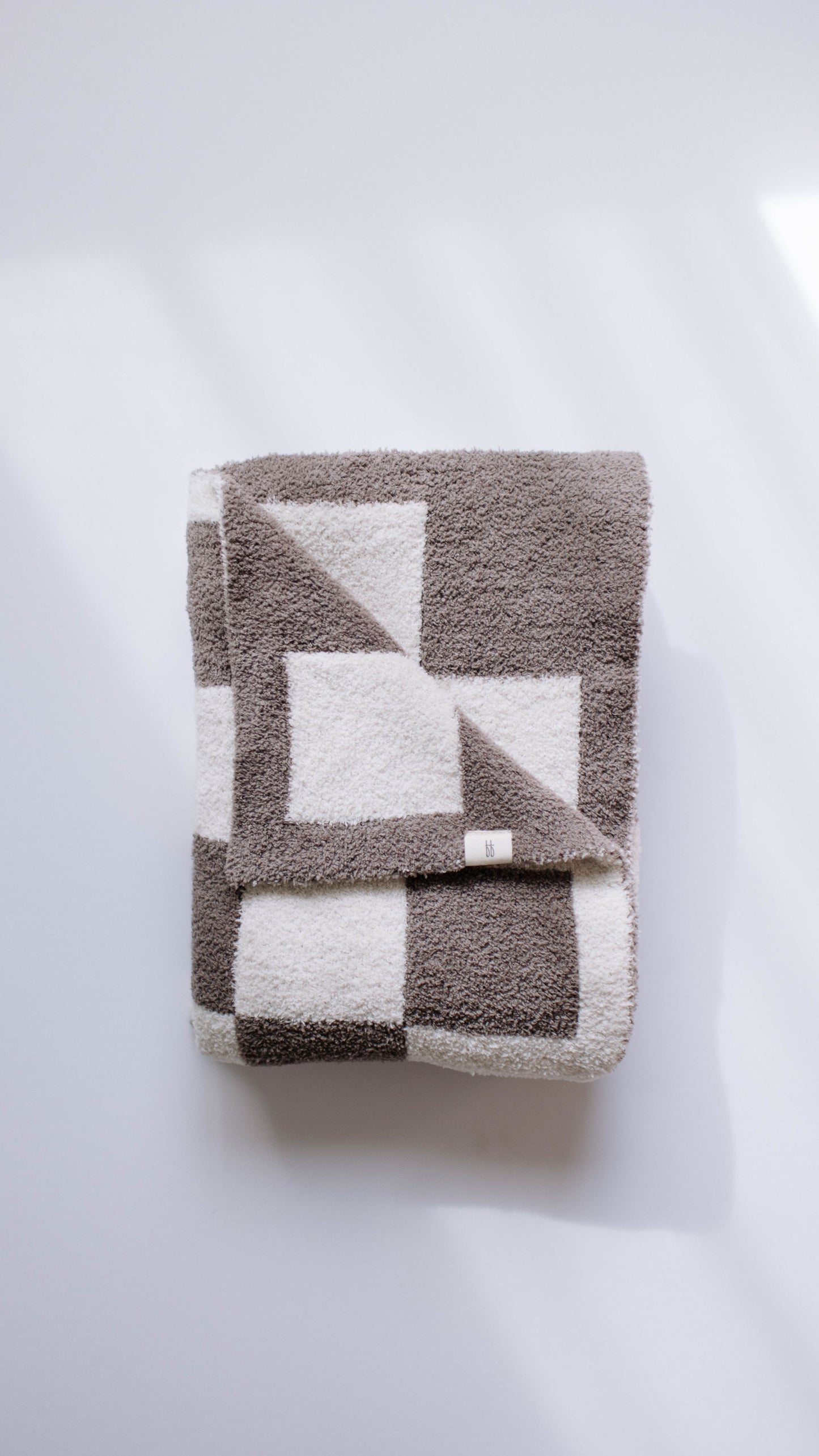 A folded, plush blanket labeled as the "Mushroom Checkerboard" by forever french baby lies on a flat white surface, its velvety-rich texture enhanced by soft light and shadows, showcasing a geometric pattern of large gray and white squares and triangles.