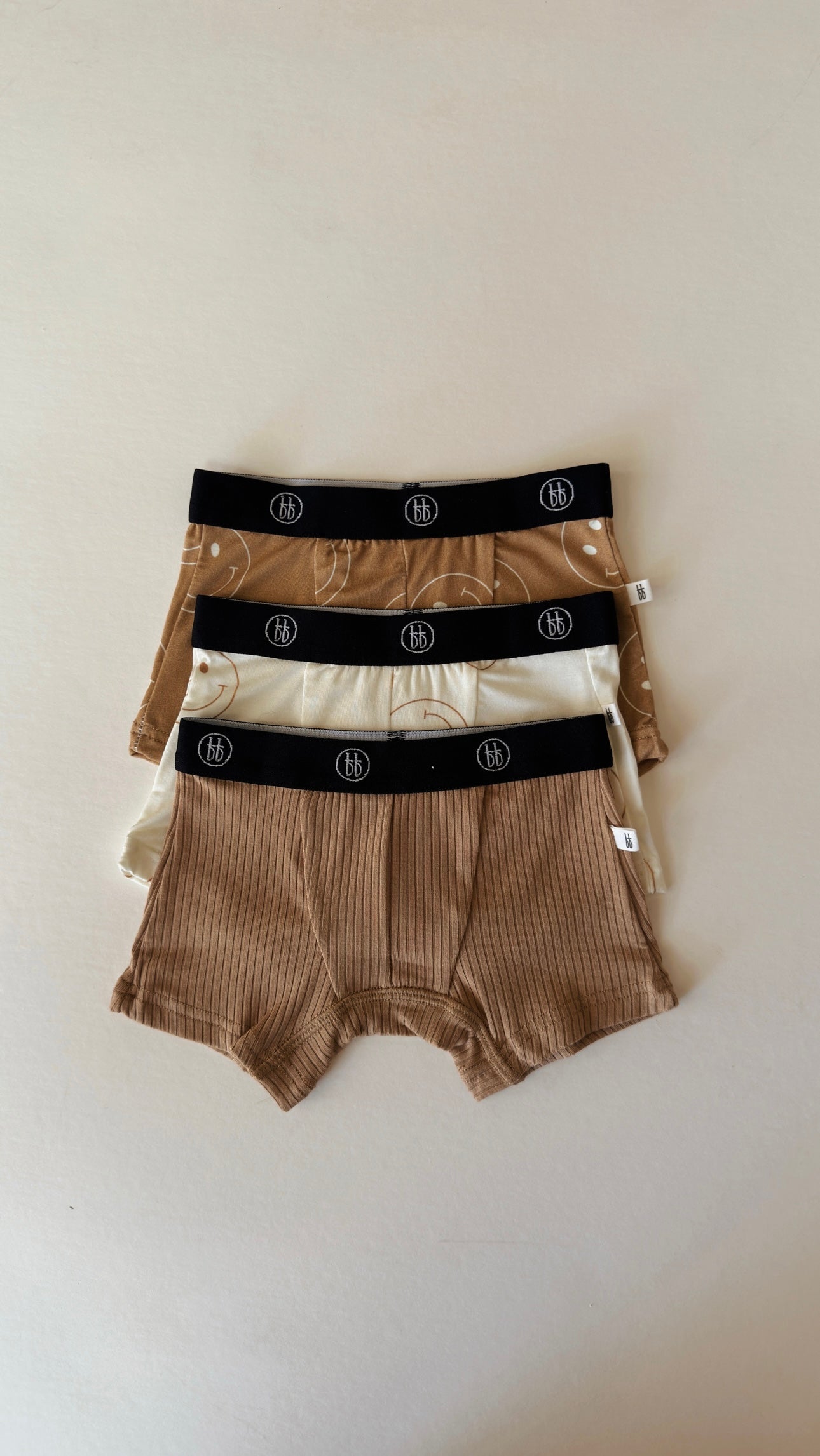 Three pairs of Bamboo Boxers from forever french baby, part of the Tan & Ivory Smile collection, are neatly arranged in a stack. These men's boxer briefs feature soothing shades of brown, tan, and beige with stylish black logo-patterned waistbands. They rest elegantly against a plain off-white surface.
