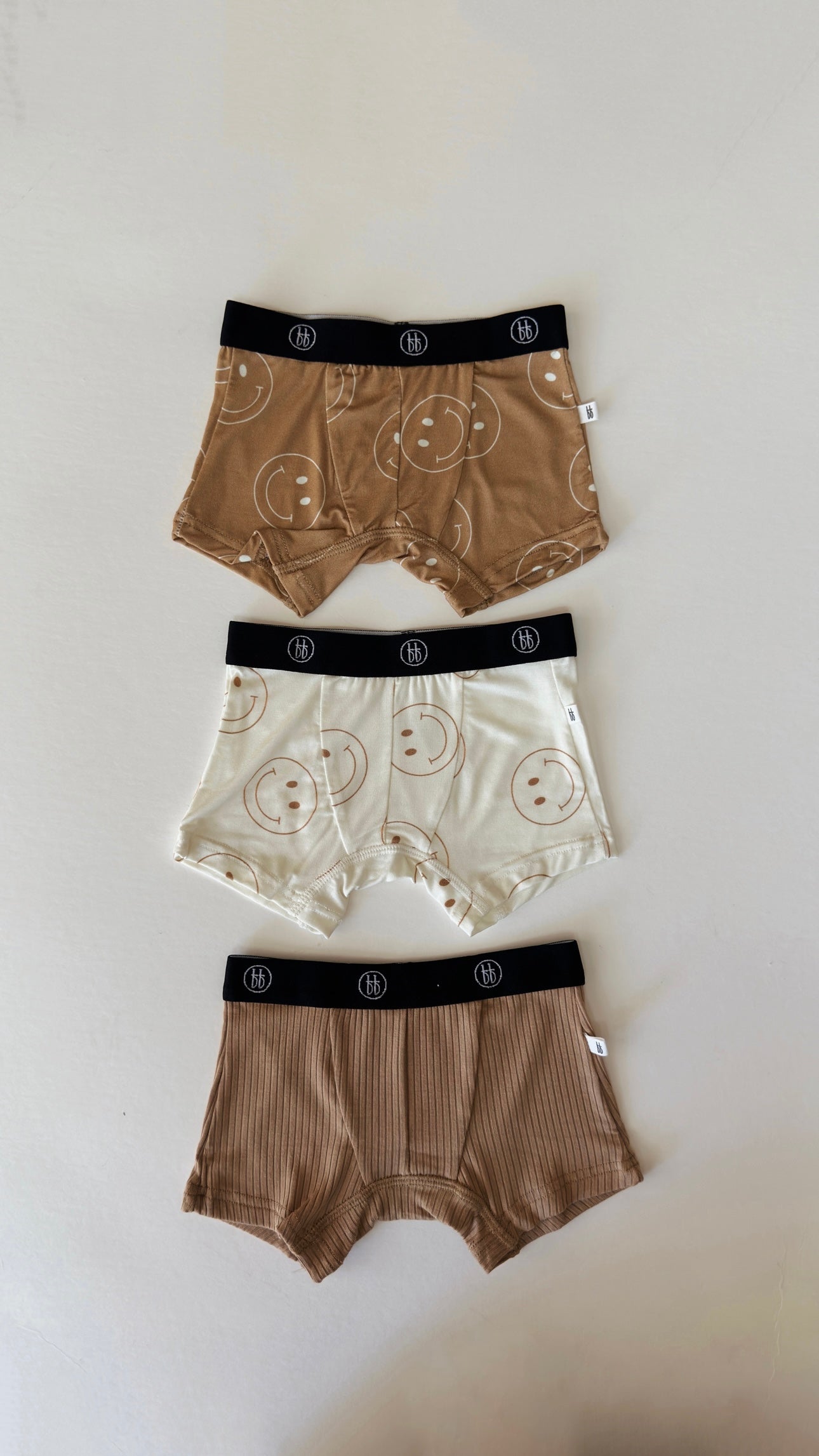 Three pairs of men's Bamboo Boxers (3 Pack) | Tan & Ivory Smile from forever french baby displayed on a flat surface. The top pair is tan with a smiley face pattern, the middle pair is ivory with a similar smiley face pattern, and the bottom pair is tan with a ribbed texture. Crafted from bamboo fabric, all have black waistbands featuring a white logo.