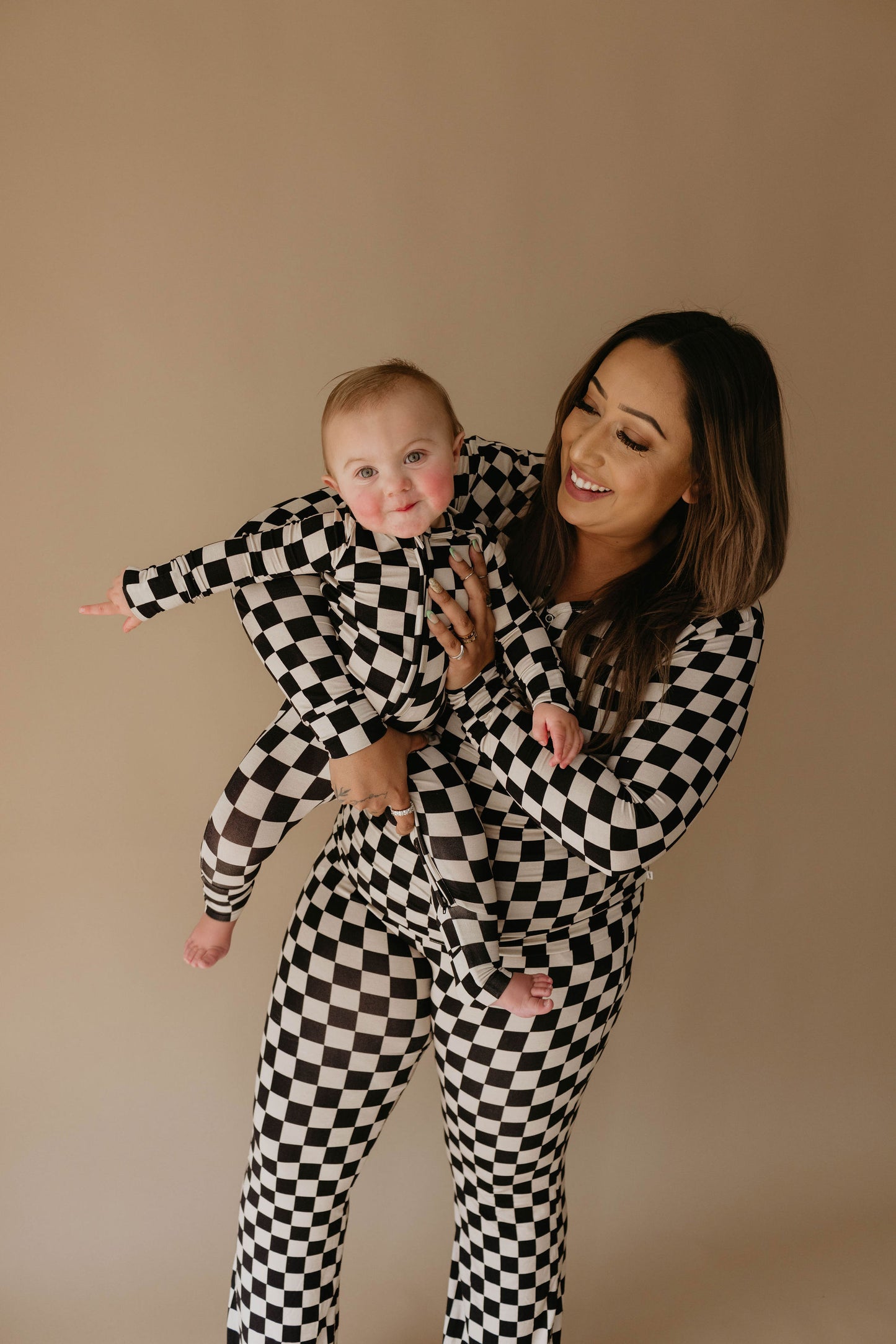 A woman with long brown hair is smiling as she holds a baby. Both are wearing forever french baby's Women's Bamboo Pajamas in the Black Checkerboard pattern, known for their softness and comfort. The baby is extending one arm outward. The background is a plain beige color.