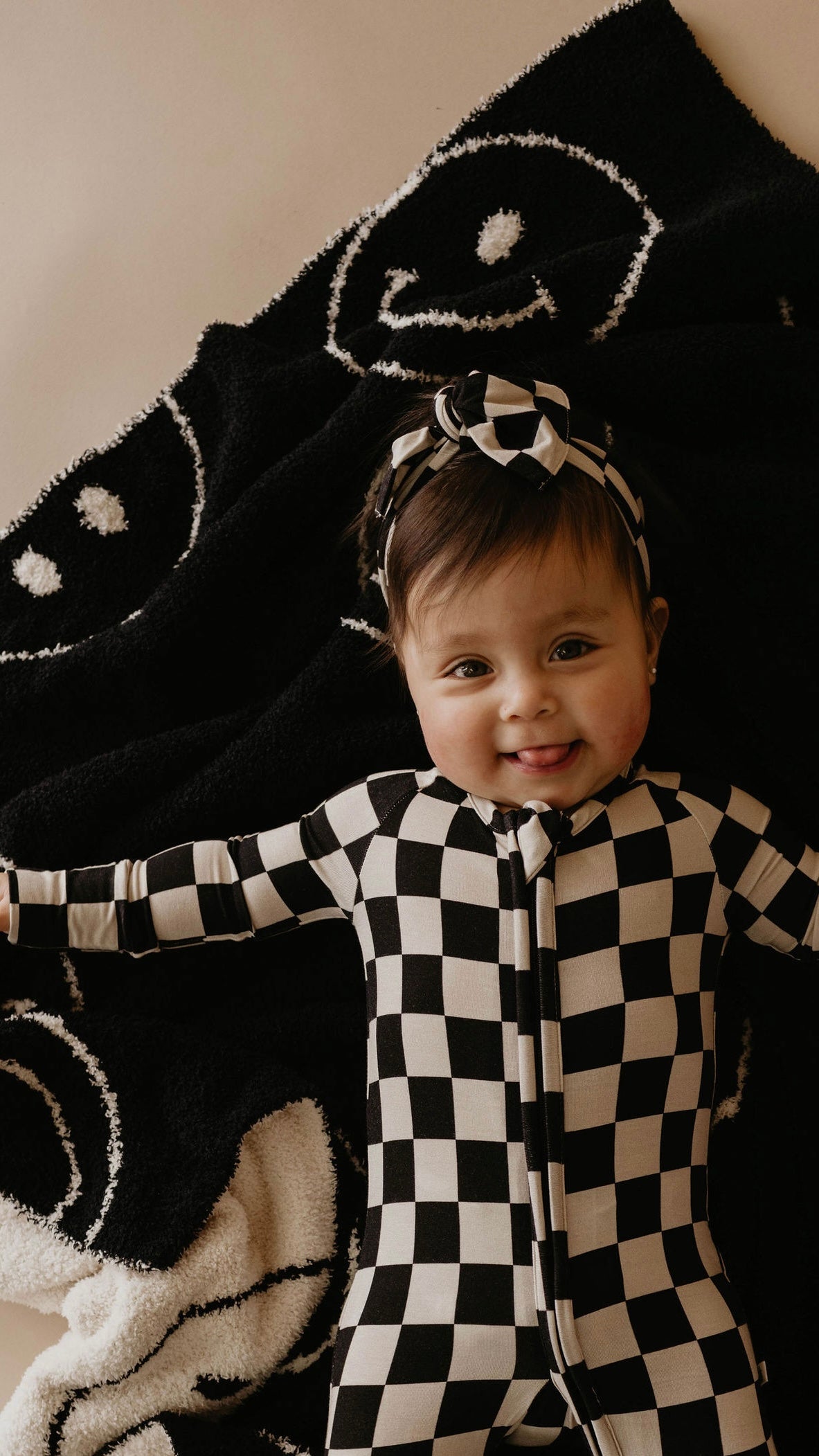 A baby wearing Bamboo Zip Pajamas in Black Checkerboard from forever french baby, along with a matching headband, is lying on a black and white blanket adorned with smiley faces. The premium pajamas feature rollover cuffs, ensuring the baby's arms stay comfortable as they stretch out and smile at the camera.