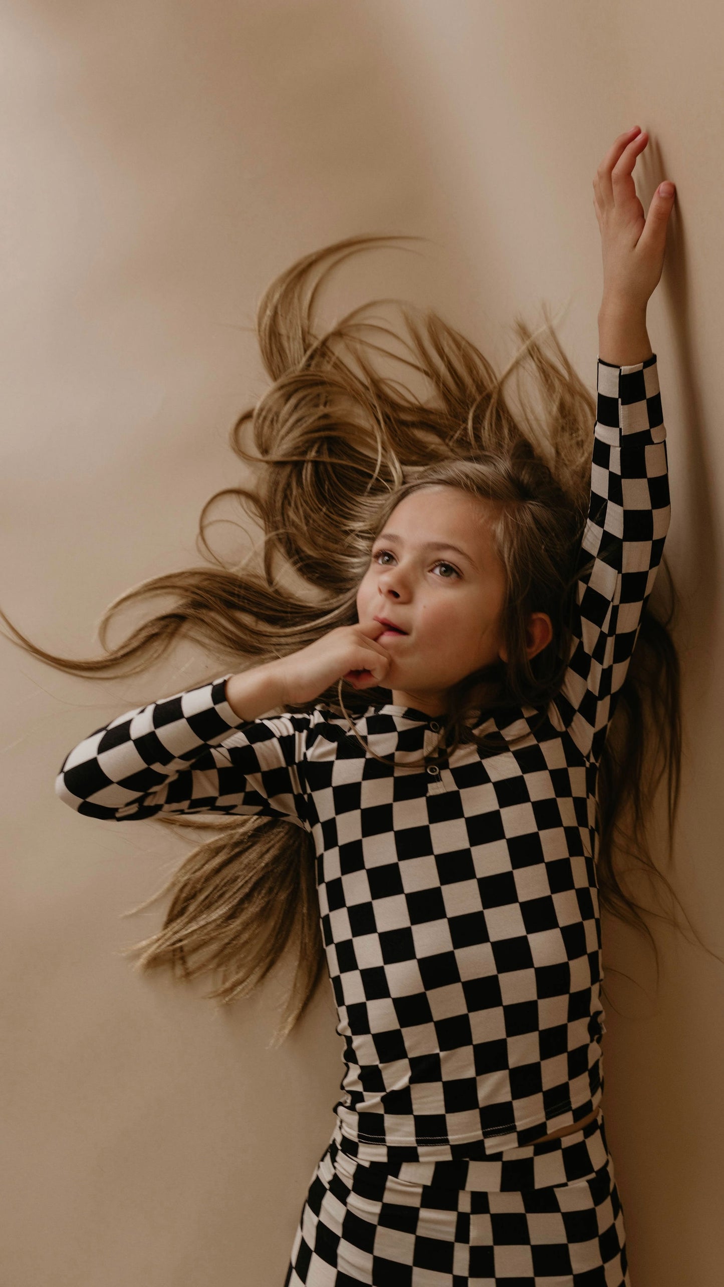 A young girl lies on a beige surface, her long hair fanning out around her head. Dressed snugly in cozy Bamboo Two Piece Pajamas | Black Checkerboard from forever french baby, her left arm is extended above her head while her right hand rests thoughtfully near her mouth. She gazes upwards.