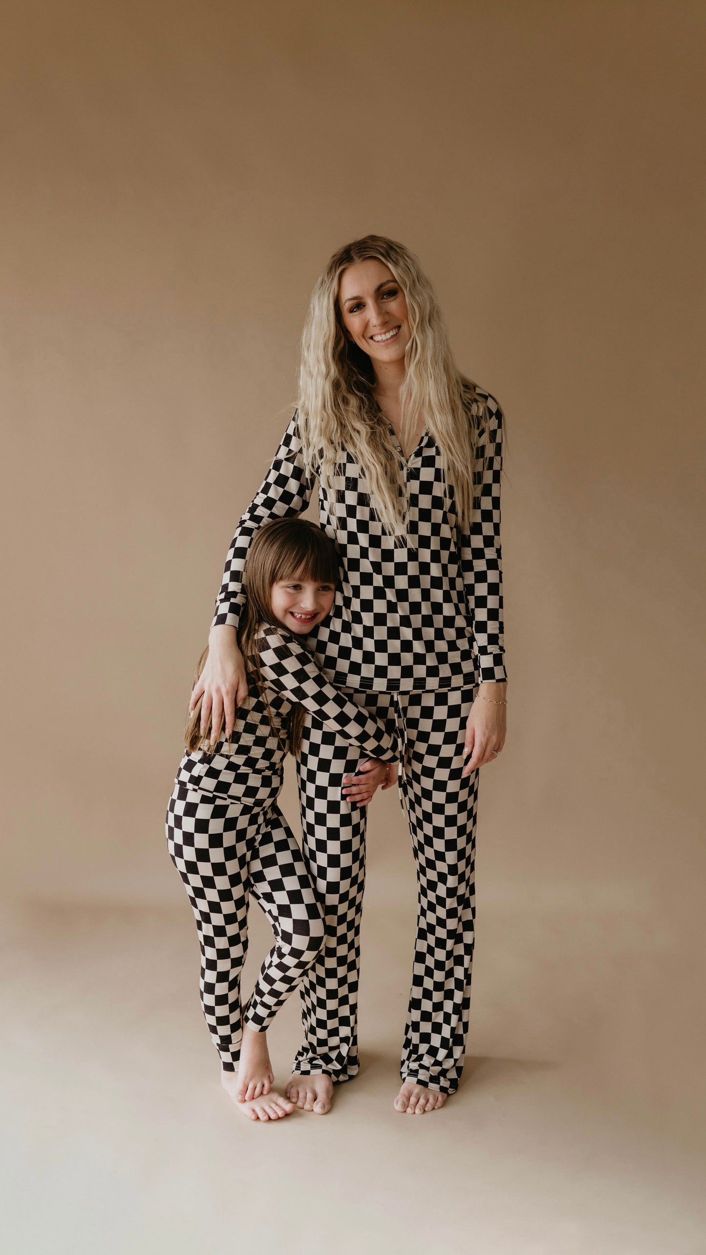 A woman and a child stand together in matching Women's Bamboo Pajamas in Black Checkerboard from forever french baby. The child hugs the woman's leg with a bright smile. They both have long hair and are barefoot, posing against a plain beige background.