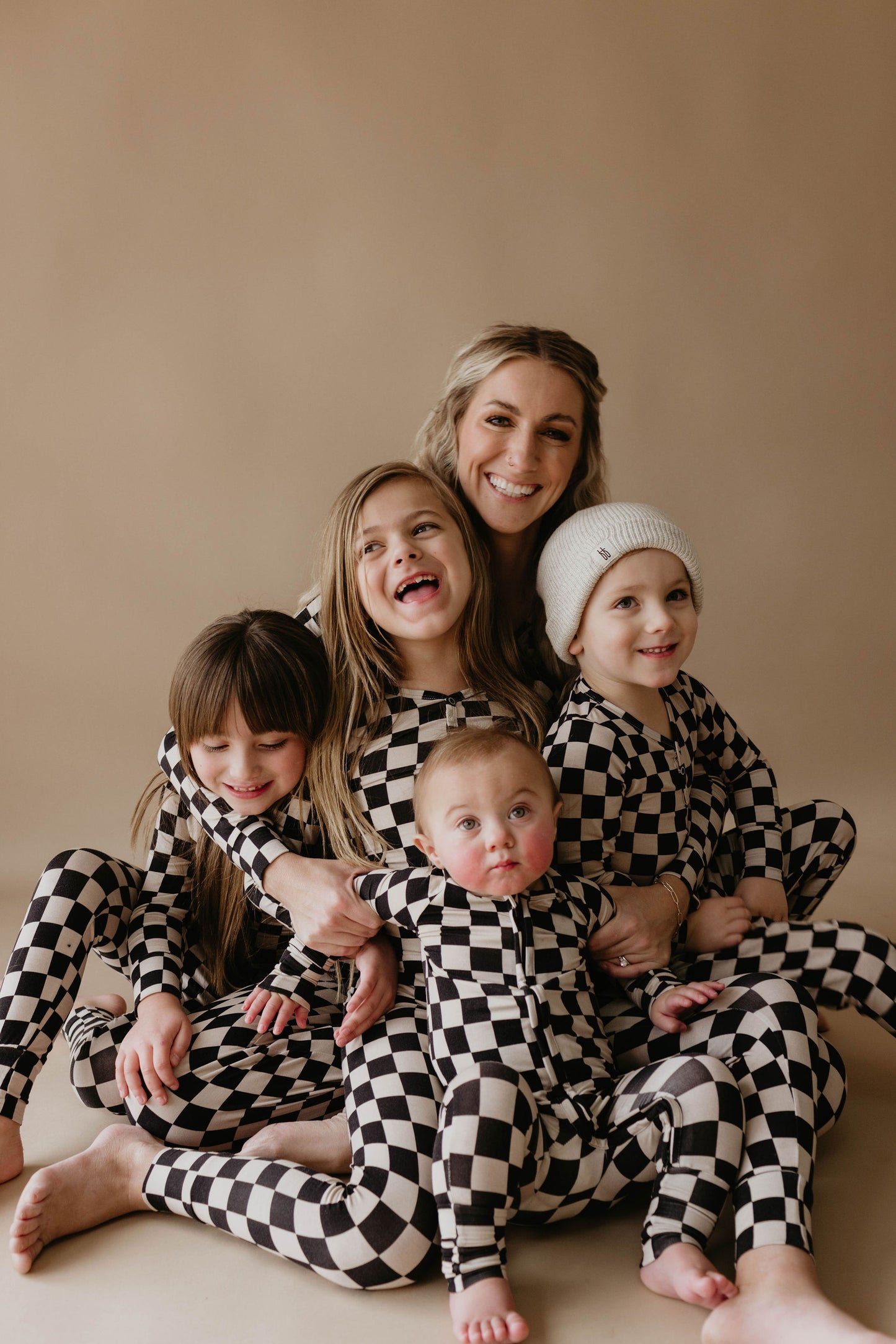 A woman sits smiling and hugging four children, all dressed in forever french baby's Women's Bamboo Pajamas in a black checkerboard pattern. The children, consisting of three girls and one boy, appear joyful, with the smallest child positioned at the front. The background is plain and neutral in color.