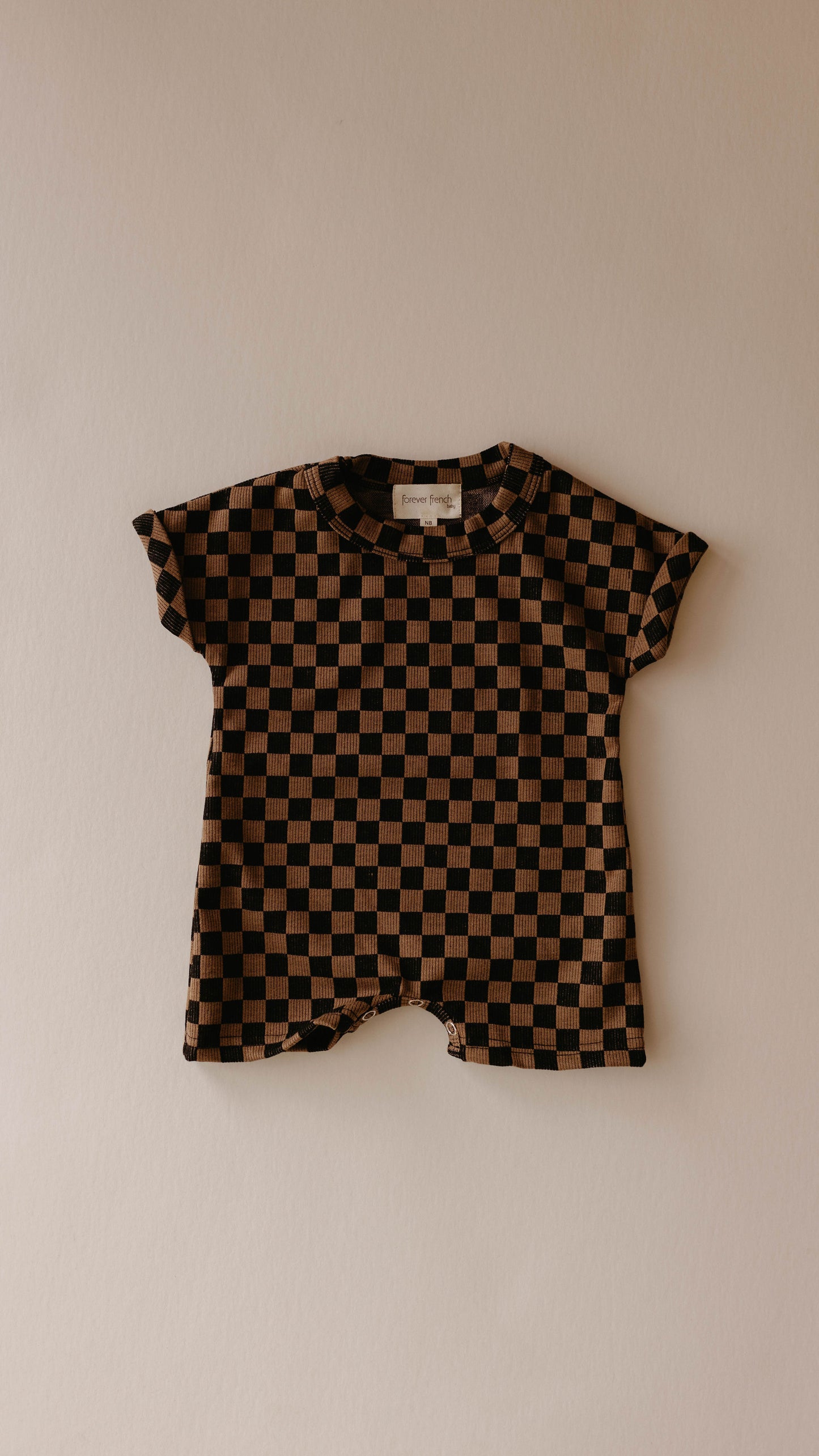 A short-sleeved, black and brown checkerboard toddler romper from forever french baby is displayed against a plain, light beige background. The Short Sleeve Ribbed Romper features a round collar and a small brand tag near the neckline.