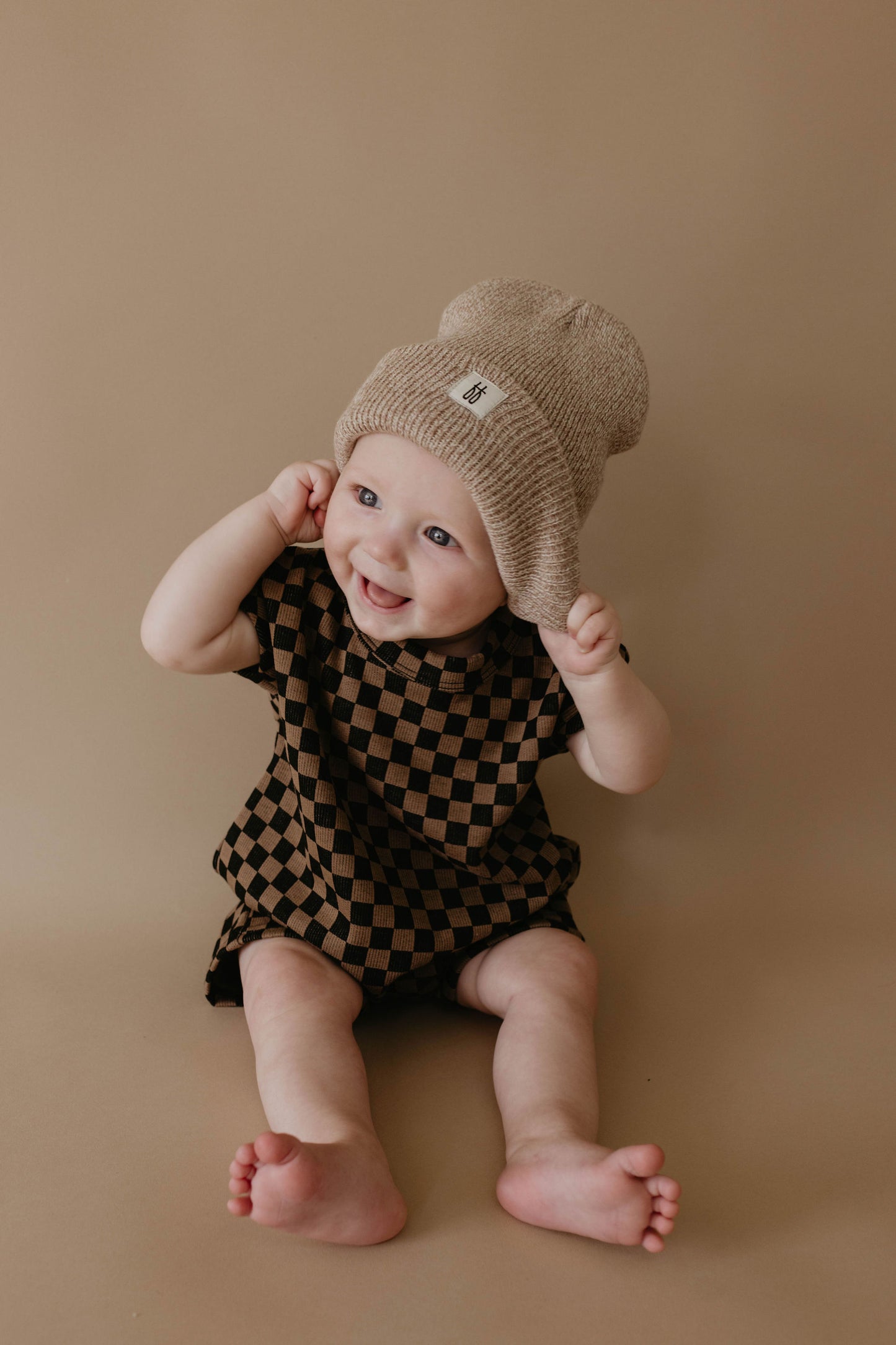 A baby sits on a beige backdrop, smiling and holding the brim of a beige knit hat on their head. The baby wears the forever french baby Short Sleeve Ribbed Romper in Black & Brown Checkerboard and is barefoot, with legs crossed in front. Perfect for easy diaper changes!