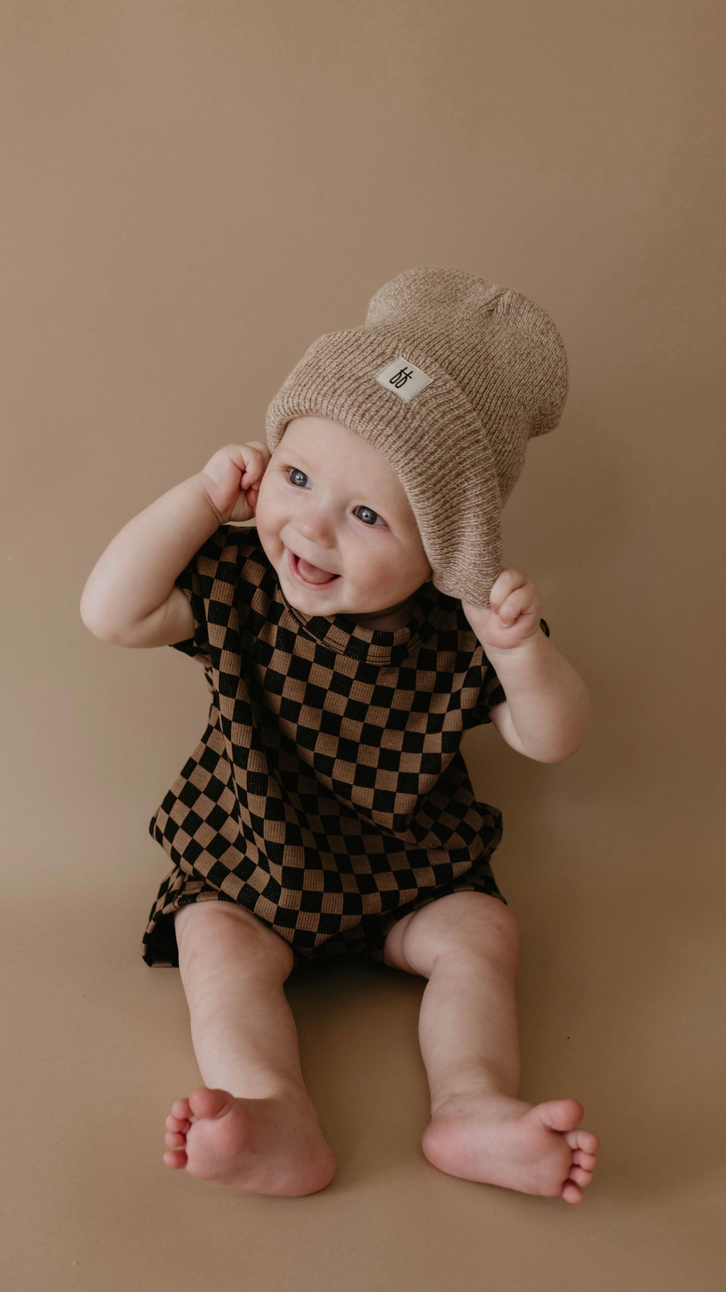 A smiling baby sits against a beige background, wearing the forever french baby Short Sleeve Ribbed Romper in Black & Brown Checkerboard and a light brown knitted hat. The romper makes diaper changes easy as the baby looks slightly off-camera, holding onto the hat with both hands.