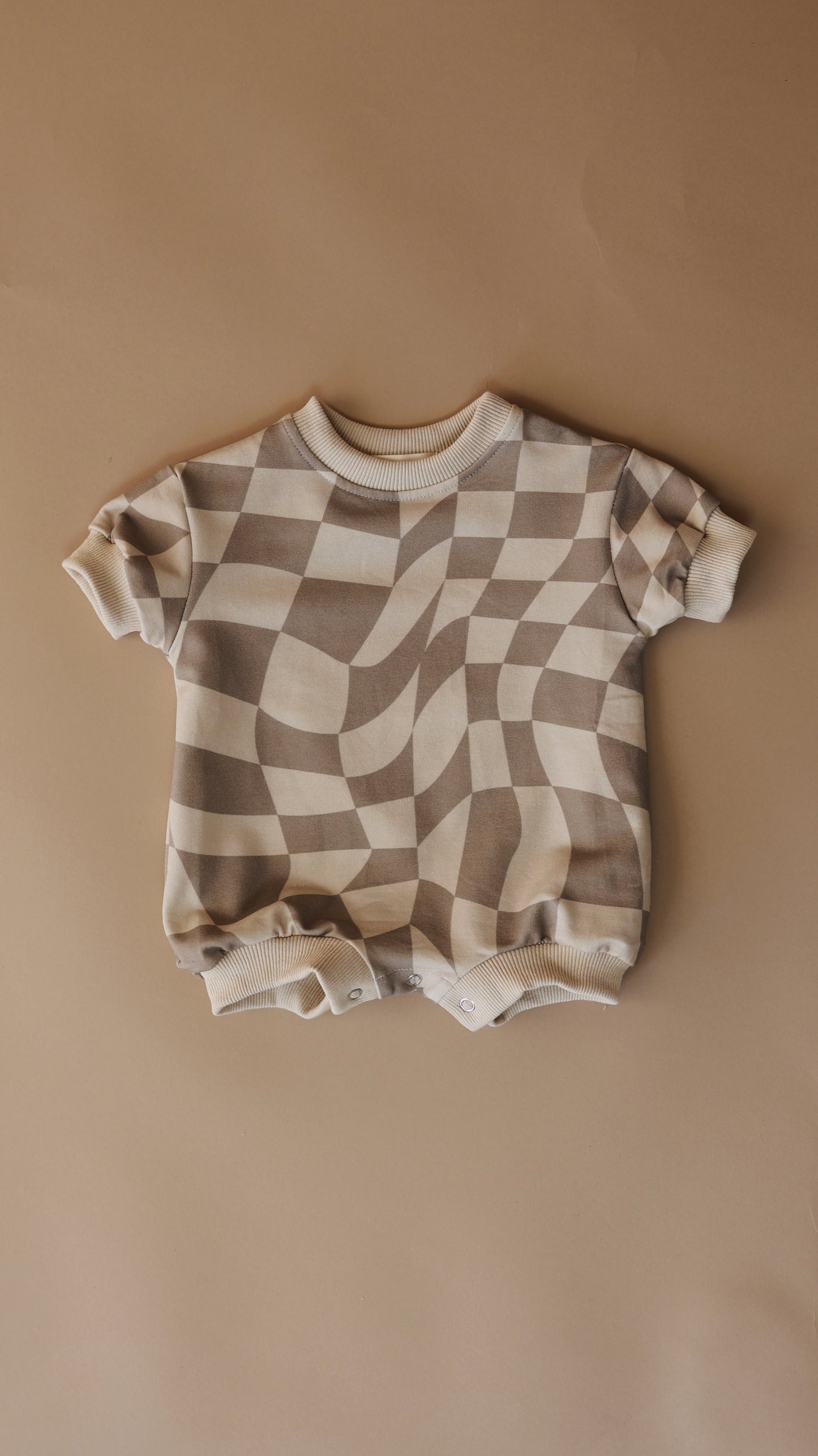 The "Short Sleeve Romper | Wavy Checkerboard" by forever french baby is a beige and cream baby romper made from organic cotton. It features short sleeves and snap buttons at the bottom and is beautifully displayed flat on a light brown background, highlighting its unique wavy checkerboard pattern.