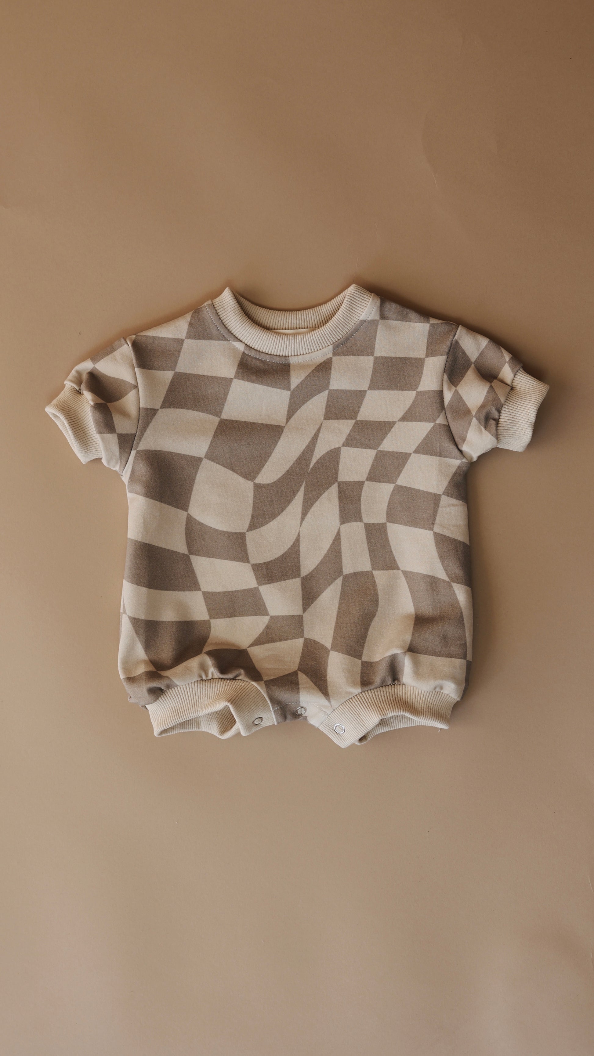 The "Short Sleeve Romper | Wavy Checkerboard" by forever french baby is a beige and cream baby romper made from organic cotton. It features short sleeves and snap buttons at the bottom and is beautifully displayed flat on a light brown background, highlighting its unique wavy checkerboard pattern.