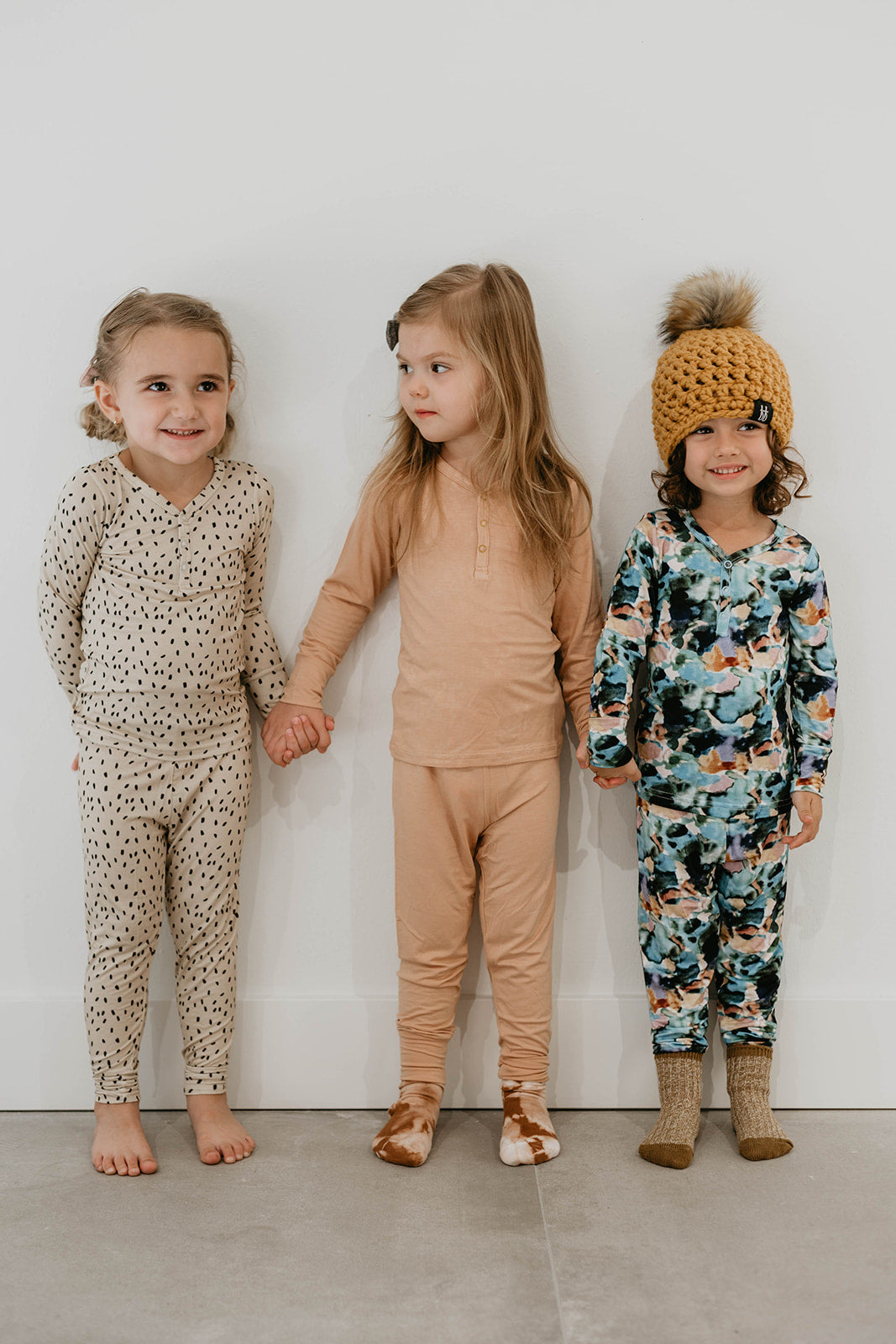Three young children stand in a row, holding hands. The child on the left wears light-colored baby pajamas with black spots. The child in the middle wears beige pajamas from forever french baby's Bamboo Zip Pajamas | Charli Print. The child on the right wears colorful pajamas and a knitted hat with a pom-pom. They are in a brightly lit room promoting Autism awareness.