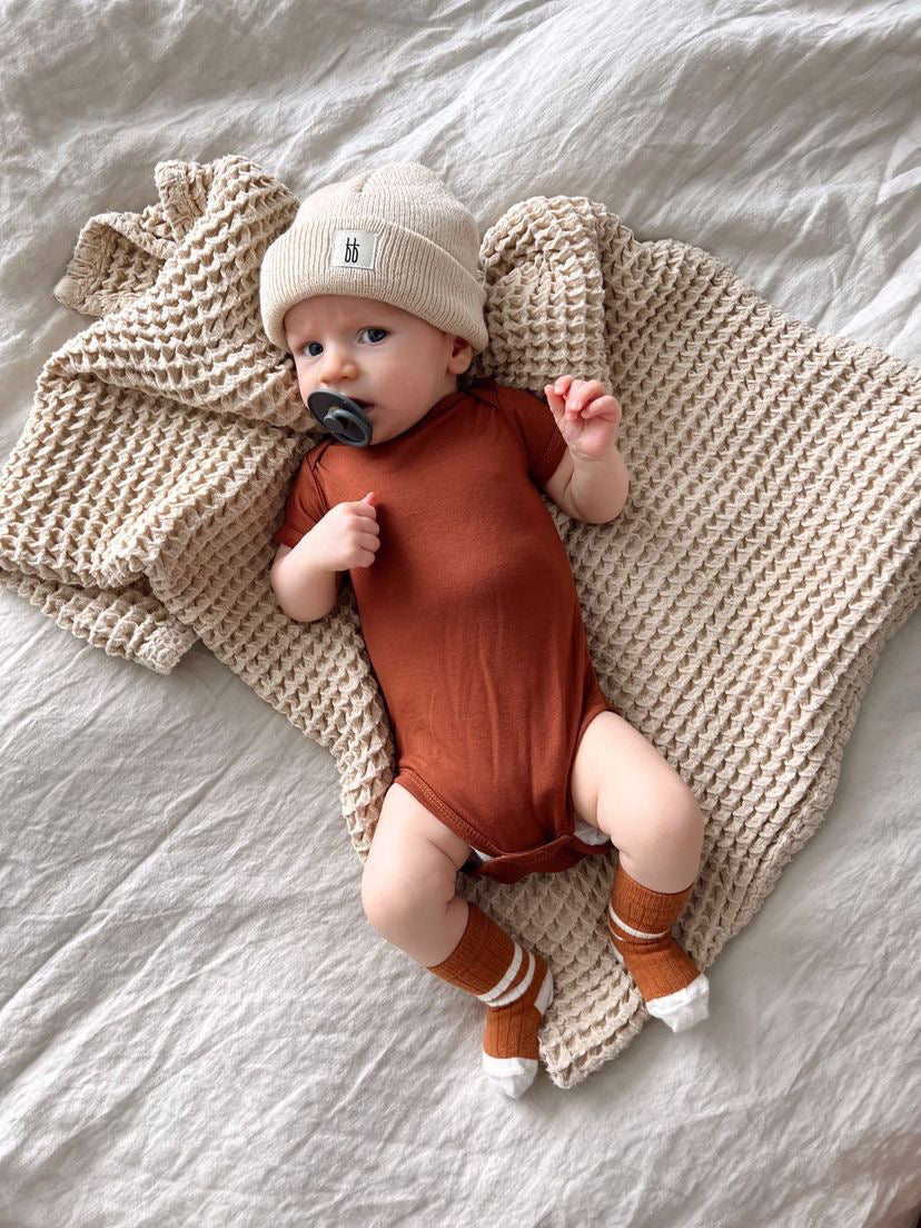 A baby wearing an oatmeal knit beanie from Forever French Baby, a rust-colored onesie, and matching socks with white tips is lying on a textured beige blanket. The baby has a grey pacifier in their mouth and is resting on a light-colored sheet from the Forever French Baby collection.