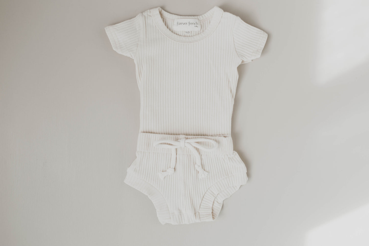 The Ribbed Set - Vanilla by forever french baby includes a white ribbed cotton onesie with short sleeves and matching shorts with a drawstring, displayed flat on a neutral background.