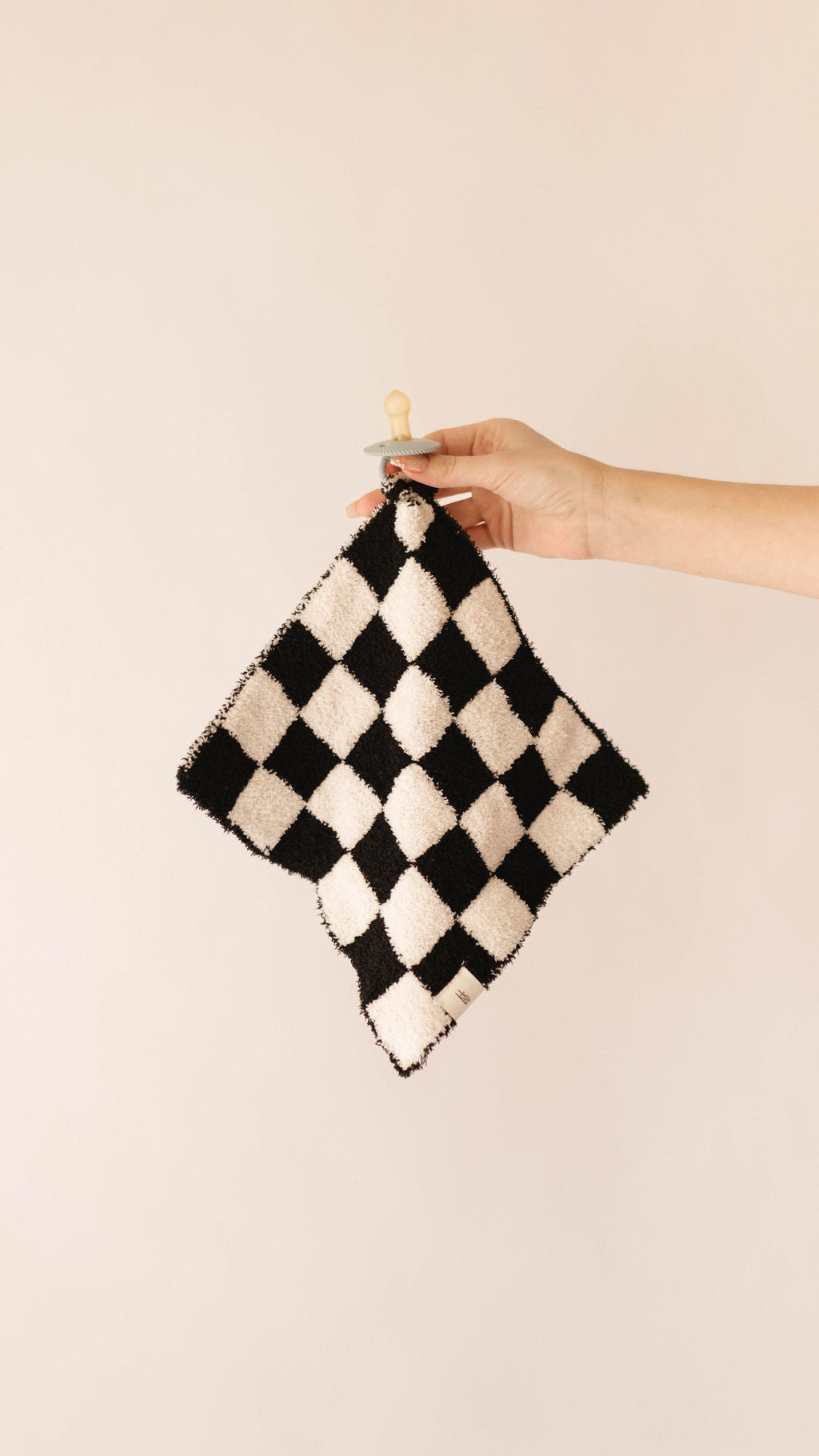 A hand holding the Lovey | Black & White Checkerboard, a small knitted blanket made from soft microfiber feather yarn with a black and white checkerboard pattern against a plain, light background. Perfect for your forever french baby collection.