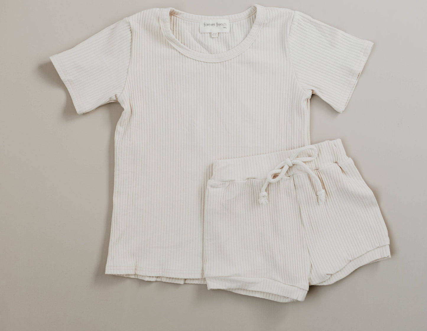 The Ribbed Set - Vanilla by forever french baby, featuring a short-sleeve beige ribbed cotton shirt and matching shorties with an elastic waistband and drawstring, is laid flat on a neutral background.