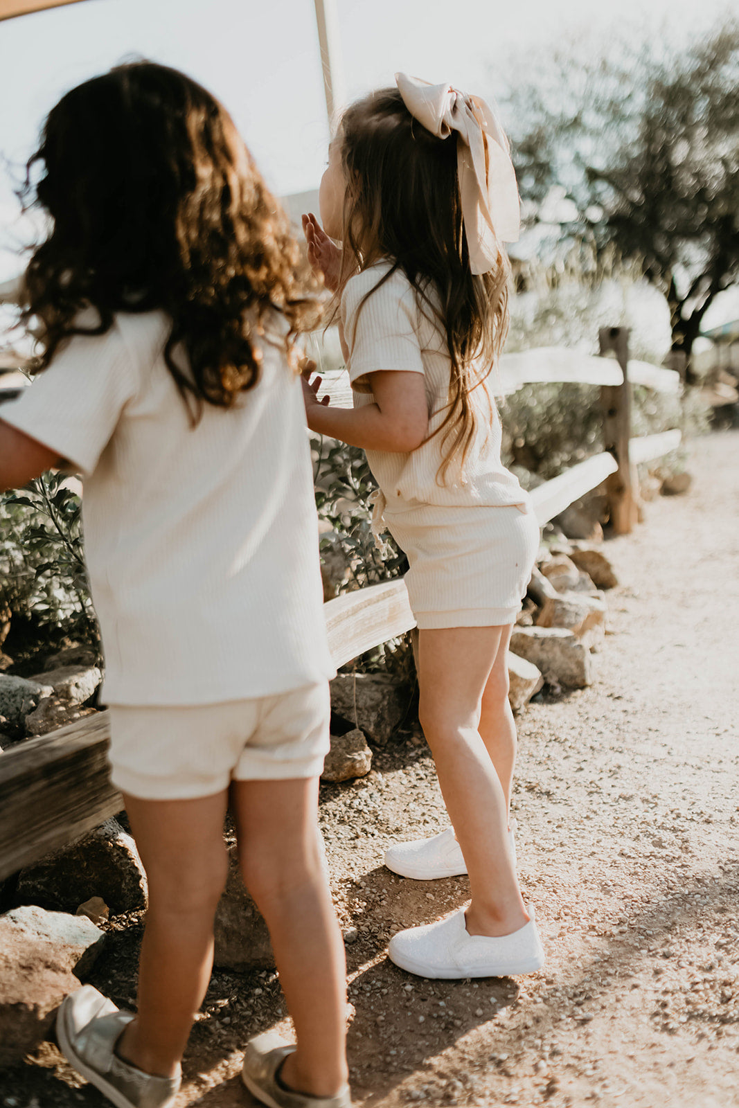 Two young girls with long hair wear the Ribbed Set - Vanilla by forever french baby. They stand outdoors near a wooden fence, surrounded by greenery and sunlight. One sports a large bow in her hair and an oversized tee, creating a warm, summery vibe.