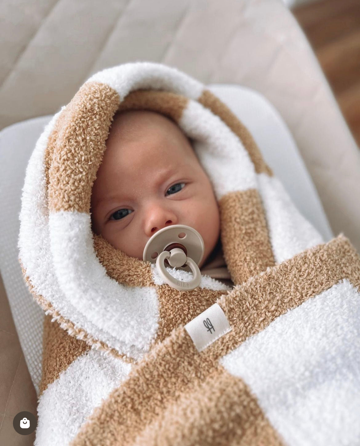 A baby wrapped snugly in a Plush Blanket | Original Checkerboard by forever french baby, lying on a light-colored surface. The baby has a pacifier in their mouth and is looking into the camera with wide eyes. The hood of the OEKO Standard 100 towel is pulled over their head, framing their face.
