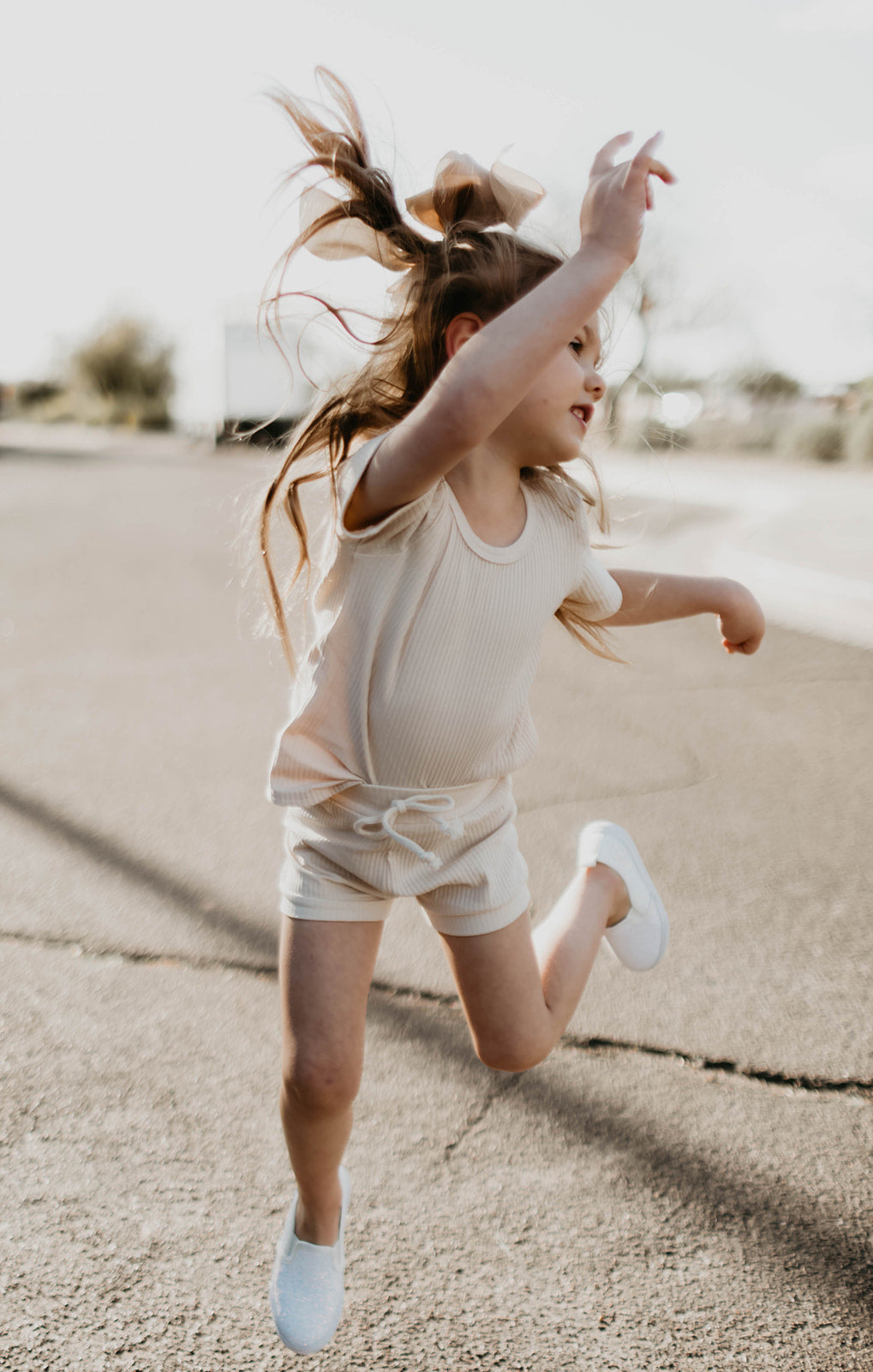 A young girl, joyfully jumping on a sunny day in her Ribbed Set - Vanilla by forever french baby, has a bow in her hair and white shoes. Her hair bounces as she raises an arm, with the blurred outdoor setting enhancing the scene.