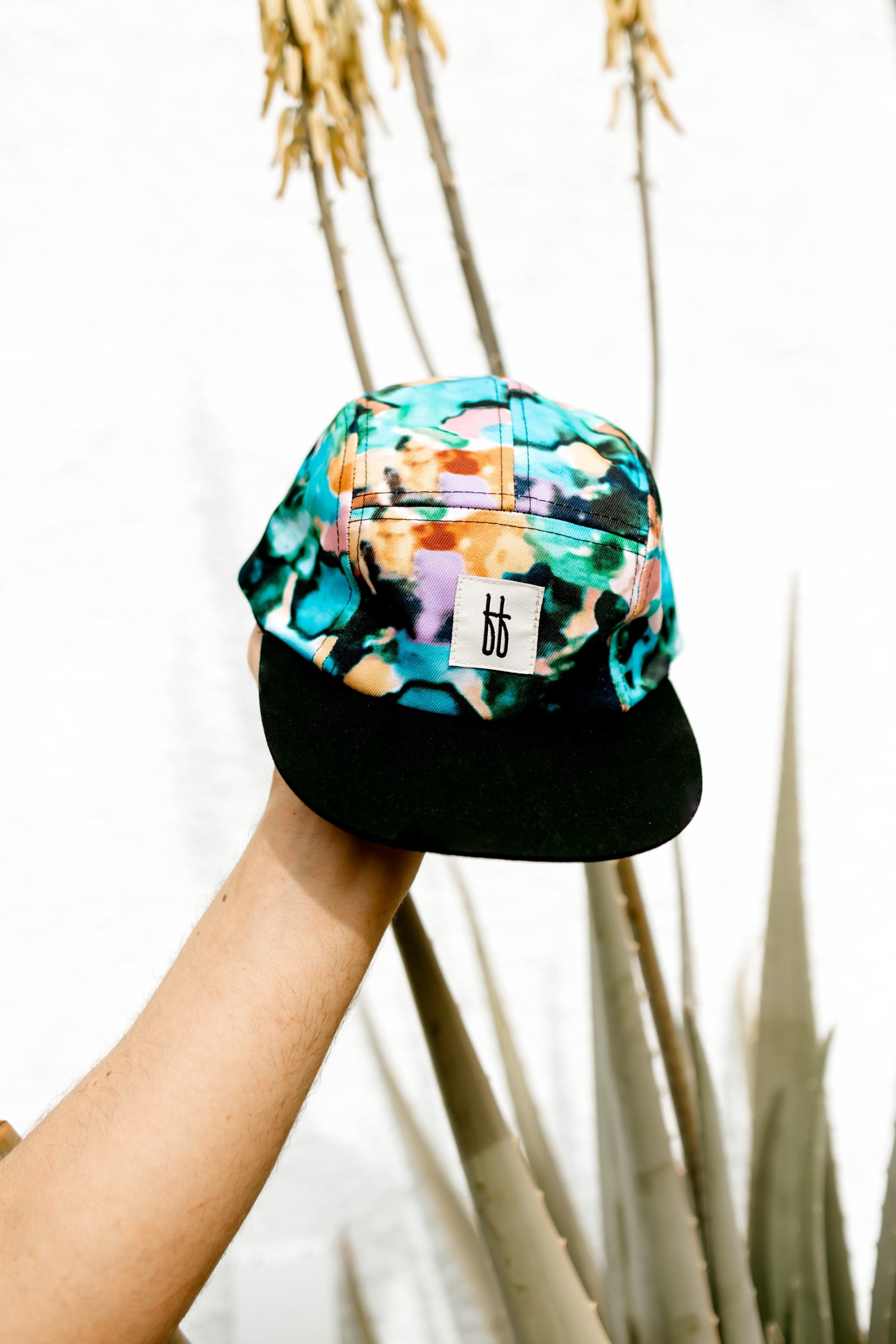 A hand is holding the Snap Back | Charli Print cap from forever french baby, featuring a colorful camouflage pattern and a black brim. This limited collection cap has a small white rectangular tag at the front with the letters "bb" printed on it. Tall, spiky plants and a white wall can be seen in the background.