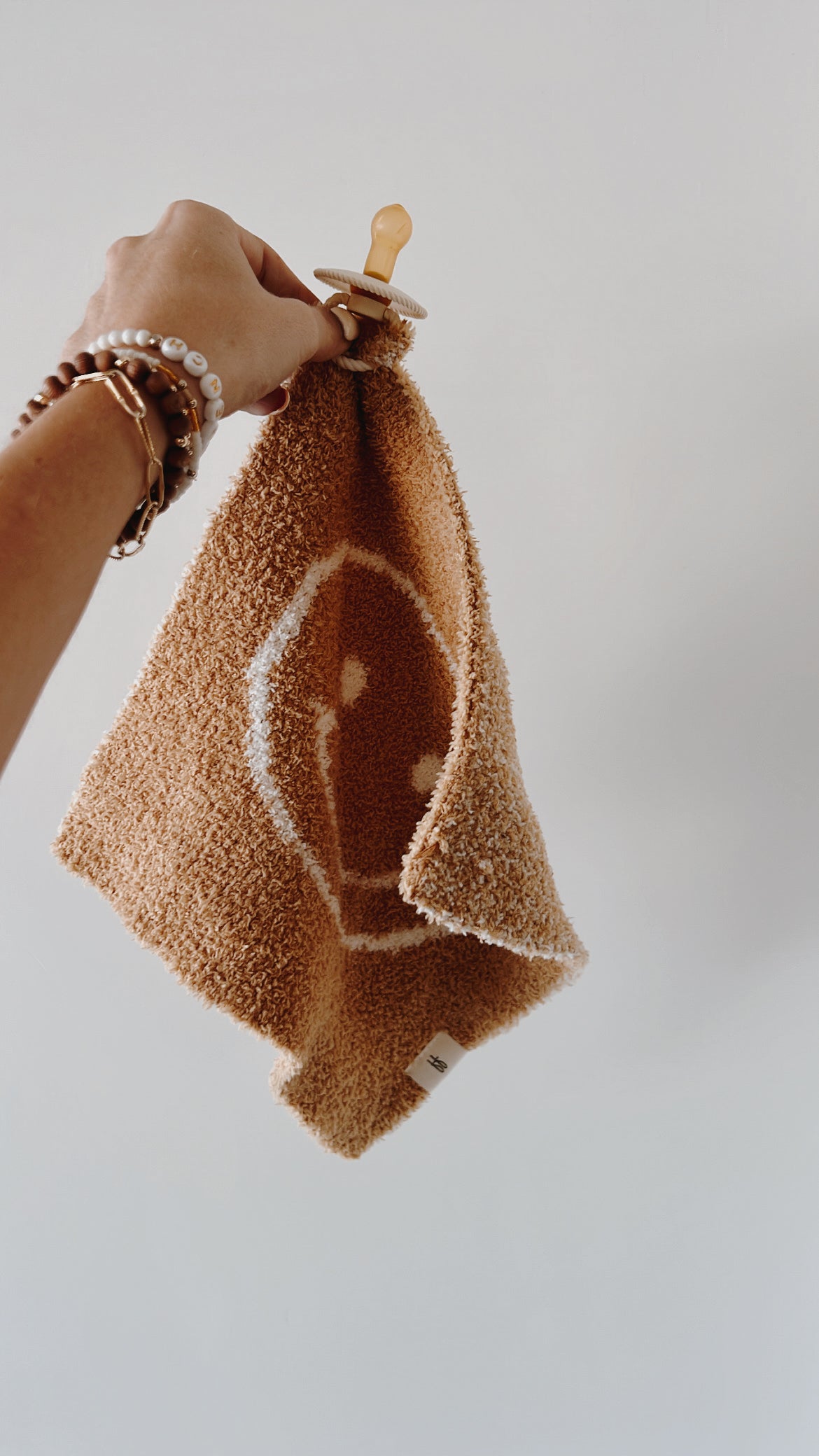 A hand holding the "Lovey | Just Smile Tan" by forever french baby, a brown microfiber feather yarn cloth adorned with a white smiley face design reminiscent of a lovey. The hand, adorned with several bracelets and an elastic hair tie, extends the cloth against a plain light-colored background.
