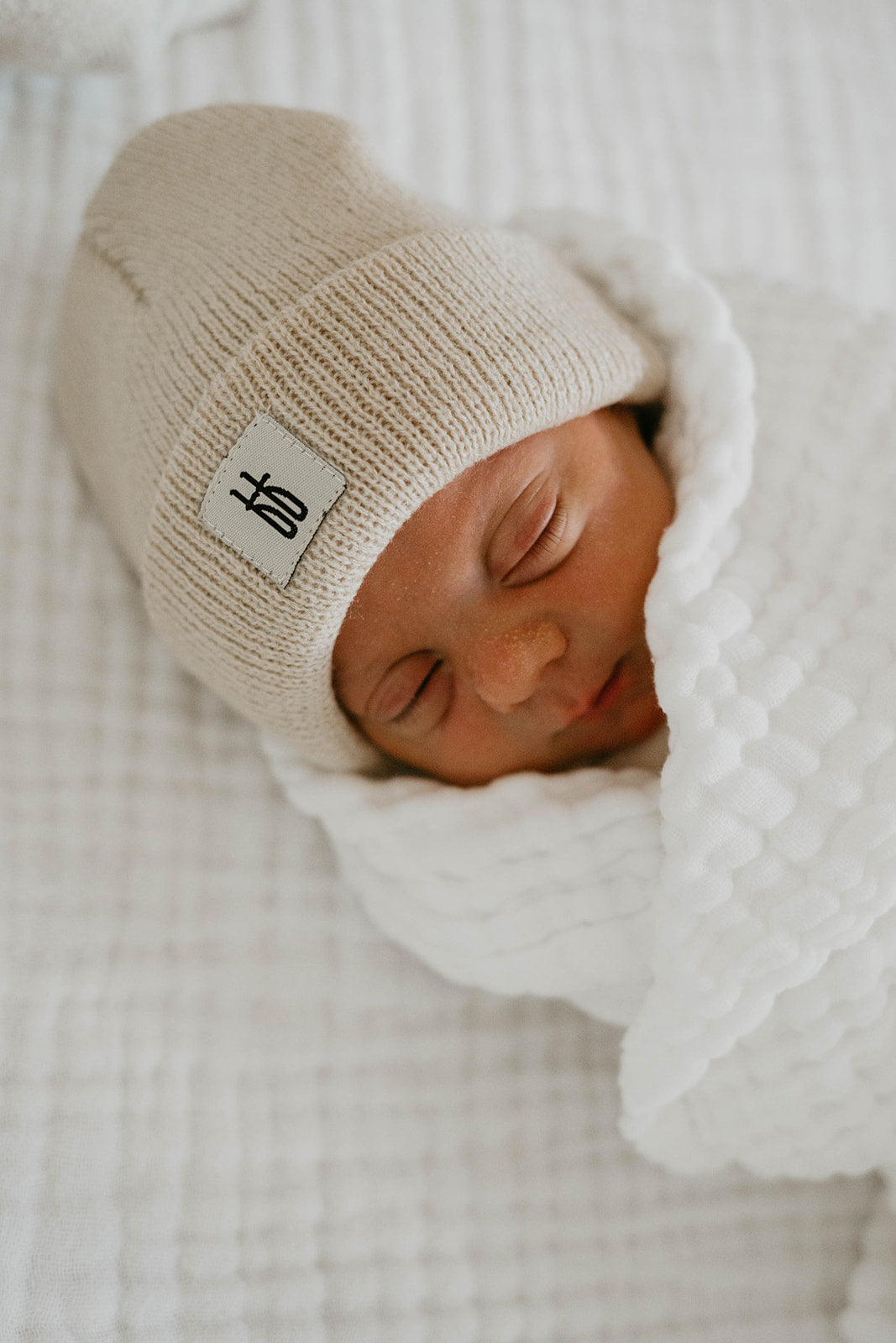 A newborn baby is swaddled in a white blanket and wearing an ff Knit Beanie | Oatmeal from forever french baby, adorned with a small label. The ribbed design complements the serene and peaceful scene as the baby sleeps soundly on a knit white surface.