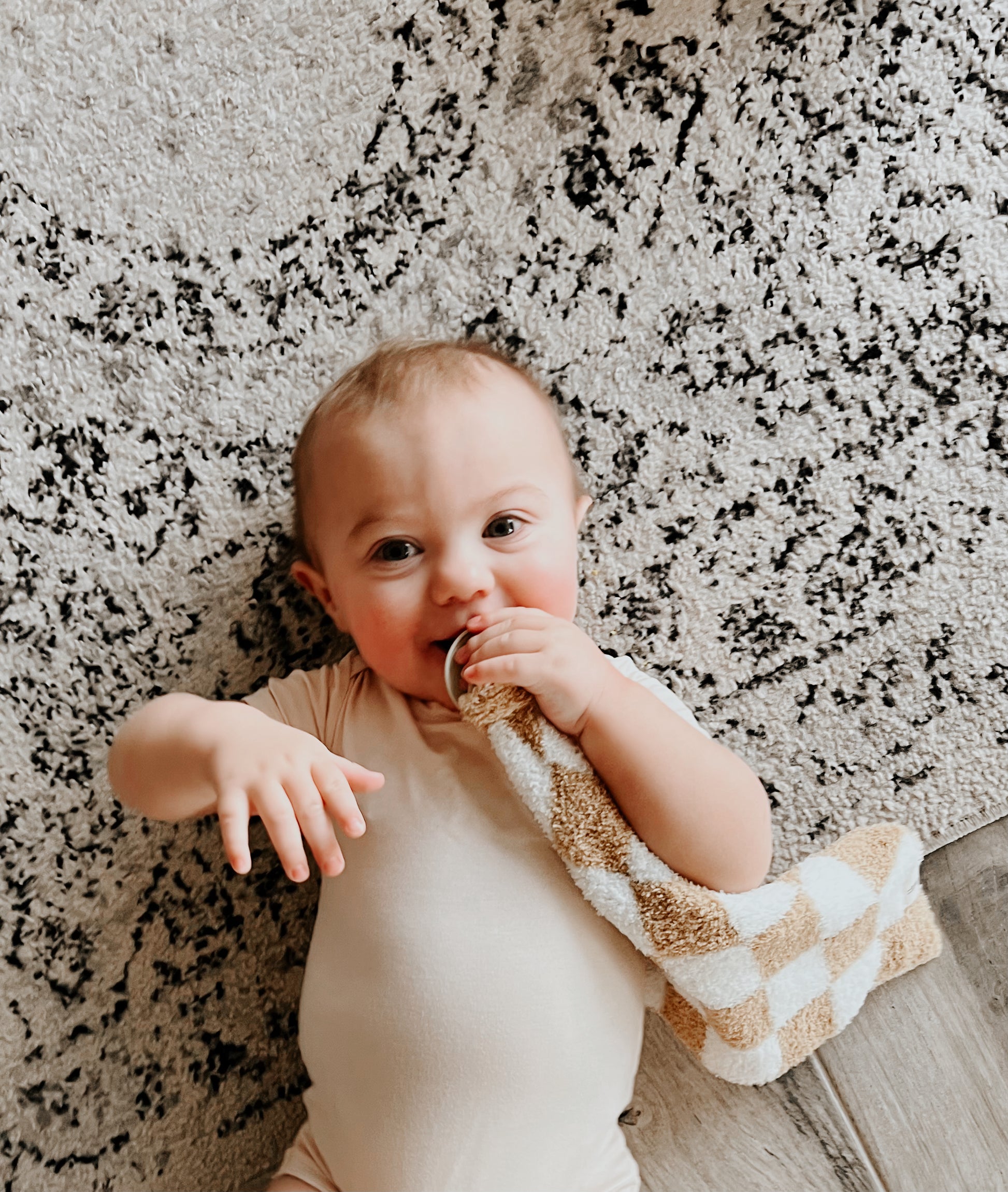A baby wearing a light-colored onesie lies on a patterned rug, holding the Lovey | Original Checkerboard from forever french baby to their mouth and looking directly at the camera with a cheerful expression. One hand is extended outward, showcasing pure joy from Forever French Baby.