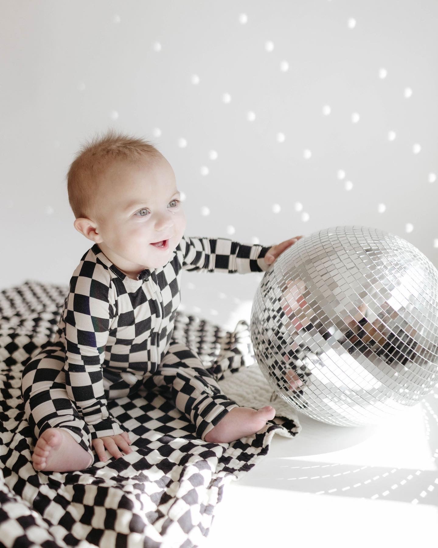 A baby wearing Bamboo Zip Pajamas in Black Checkerboard from forever french baby sits on a matching patterned blanket. The baby is smiling and reaching out to touch a large, reflective disco ball placed in front of them. The background is light, with scattered light reflections from the disco ball.