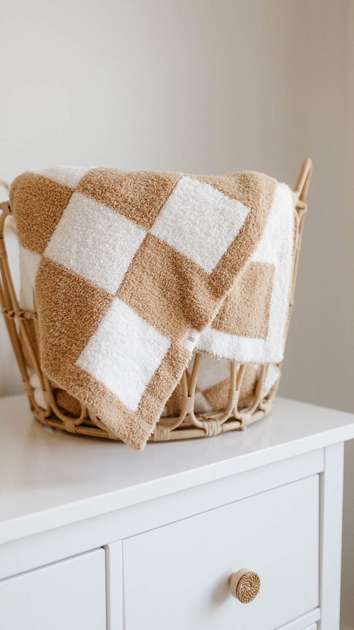 A wicker basket with a forever french baby Plush Blanket | Original Checkerboard draped over it sits on top of a white dresser with a single drawer. The scene is minimalist and cozy, with neutral tones and a calming vibe.